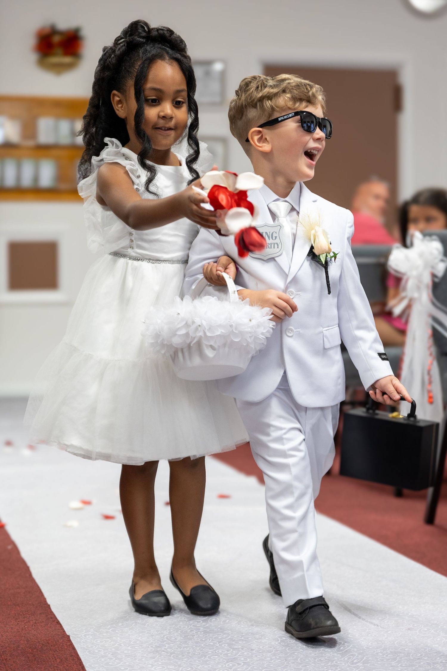 Adorable flower girl and ring bearer in white outfits walking down wedding ceremony aisle together.
