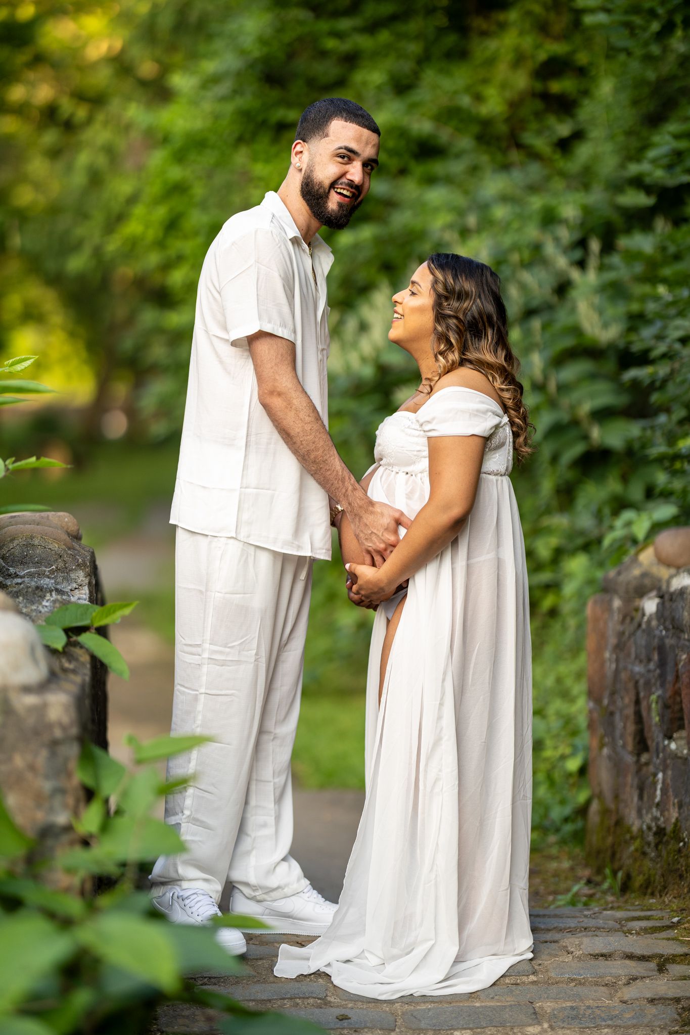 Happy couple in matching white outfits holding hands during outdoor maternity photo session.
