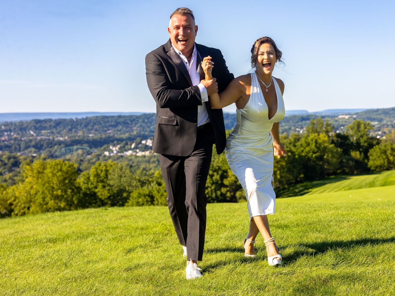 Happy couple running on a lush green hill with a scenic town view on a sunny day.