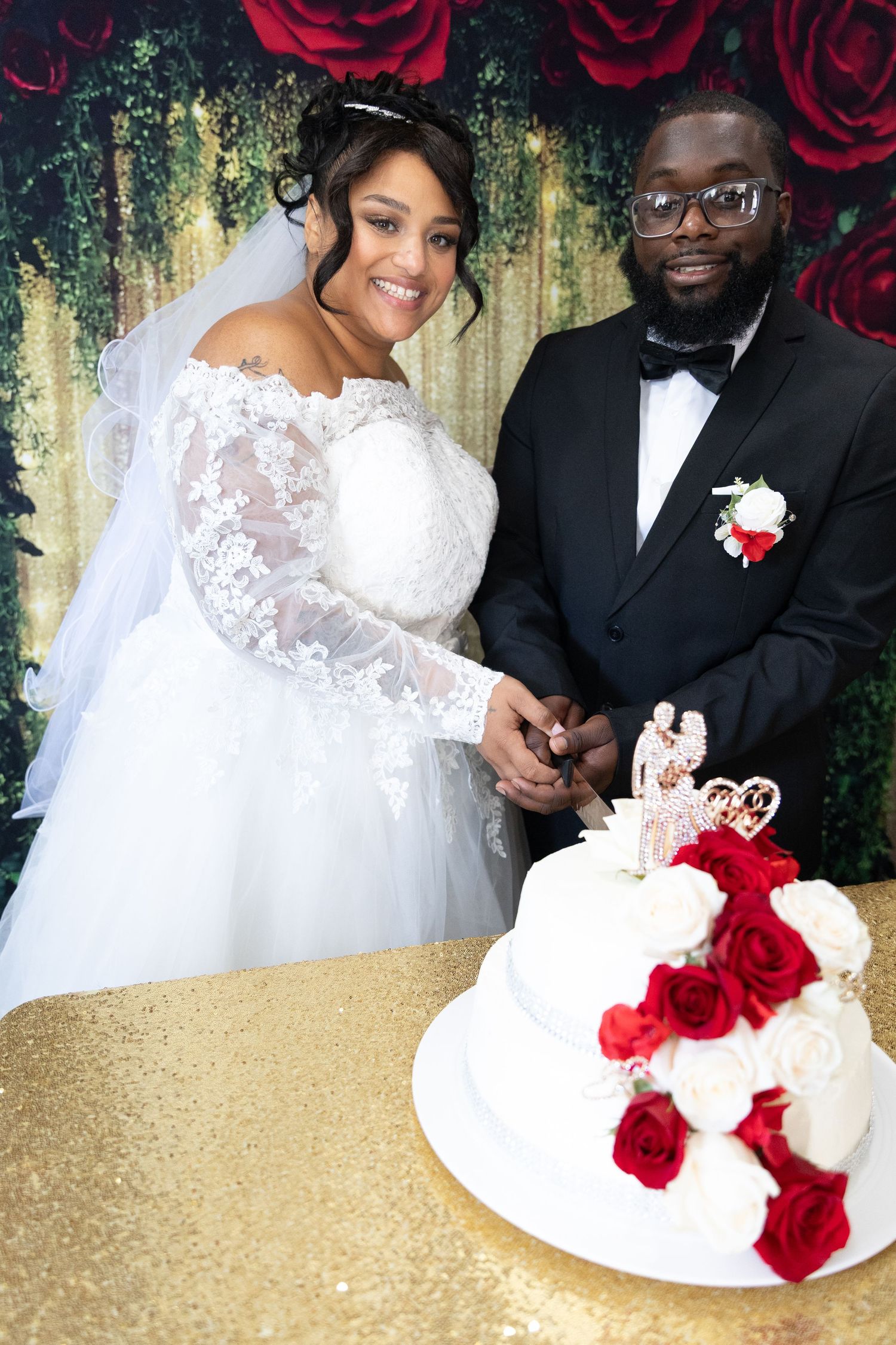 Newlywed couple cutting elegant white wedding cake decorated with red roses at reception.