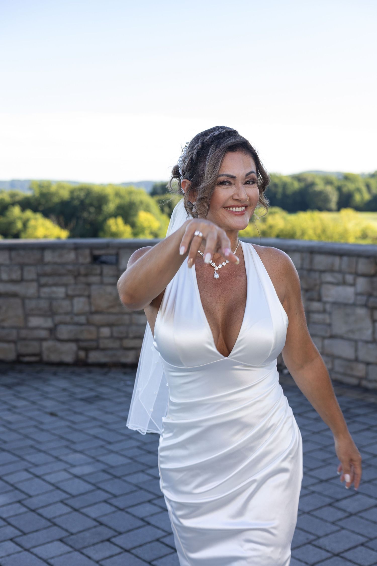 Smiling bride in white halter gown with veil extends ring hand outdoors on stone patio with trees in background.