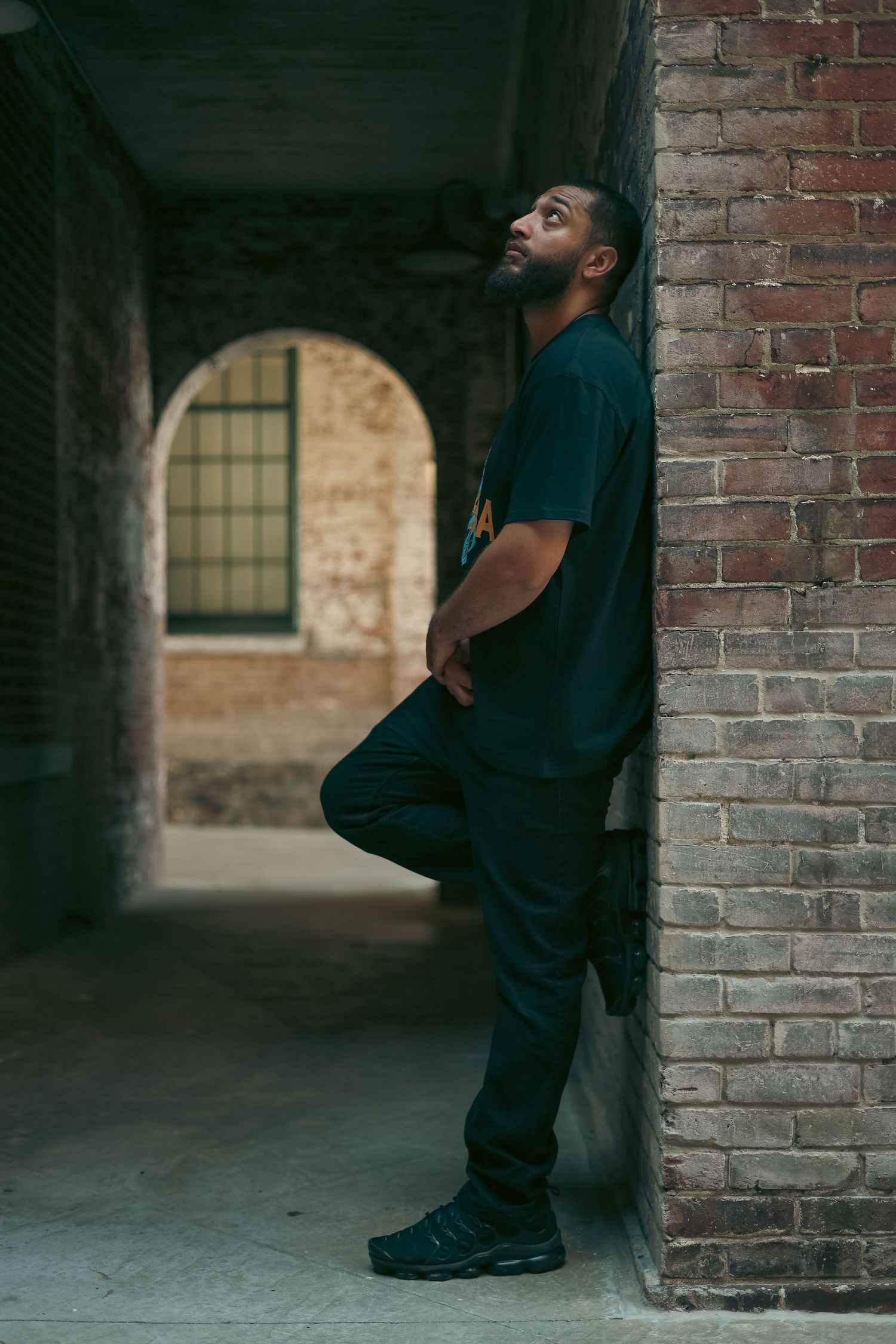 Young man in dark clothing leaning against brick wall in urban alleyway looking upward.