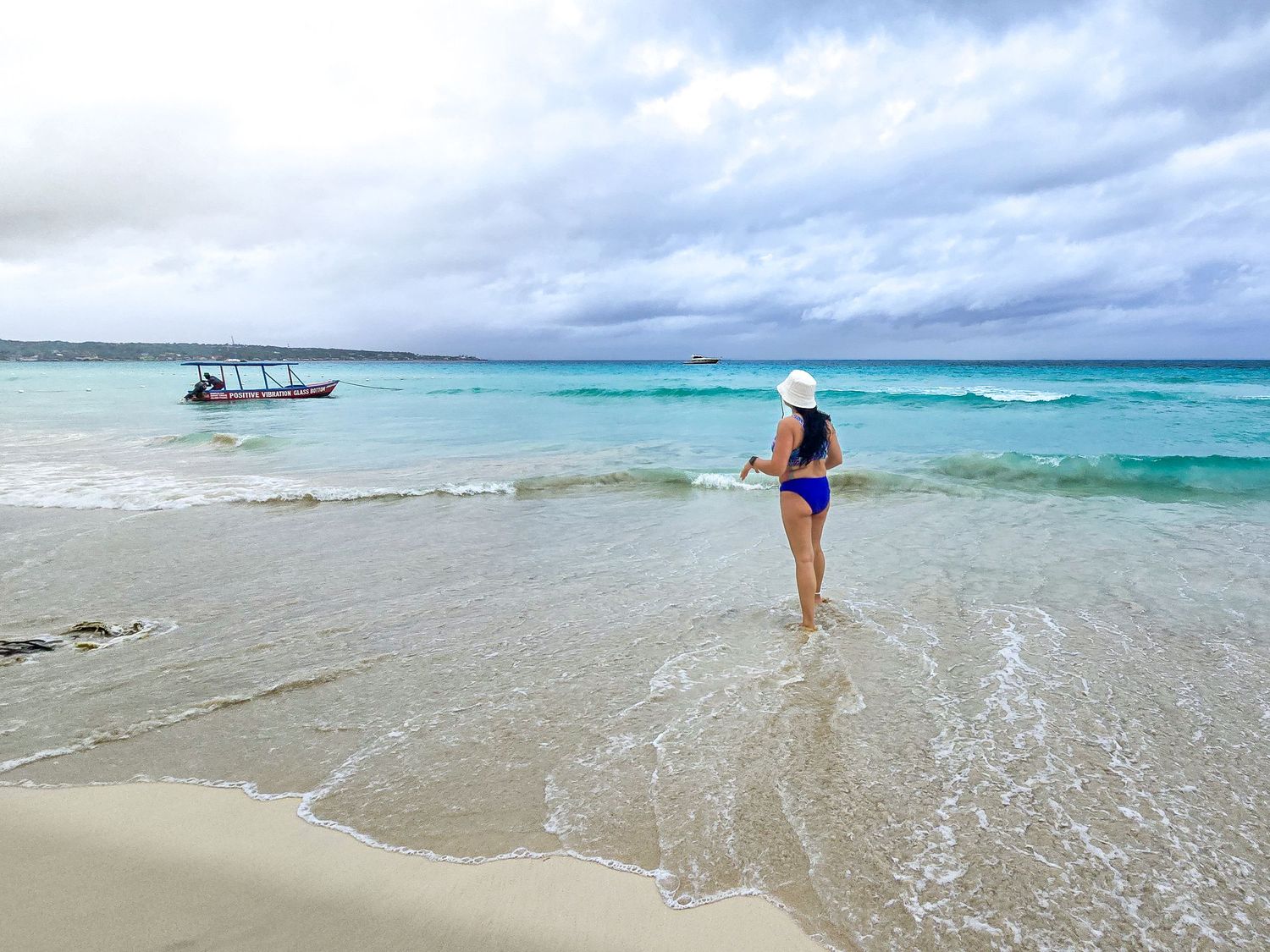 Woman in blue bikini and white hat standing at edge of turquoise tropical beach waters.