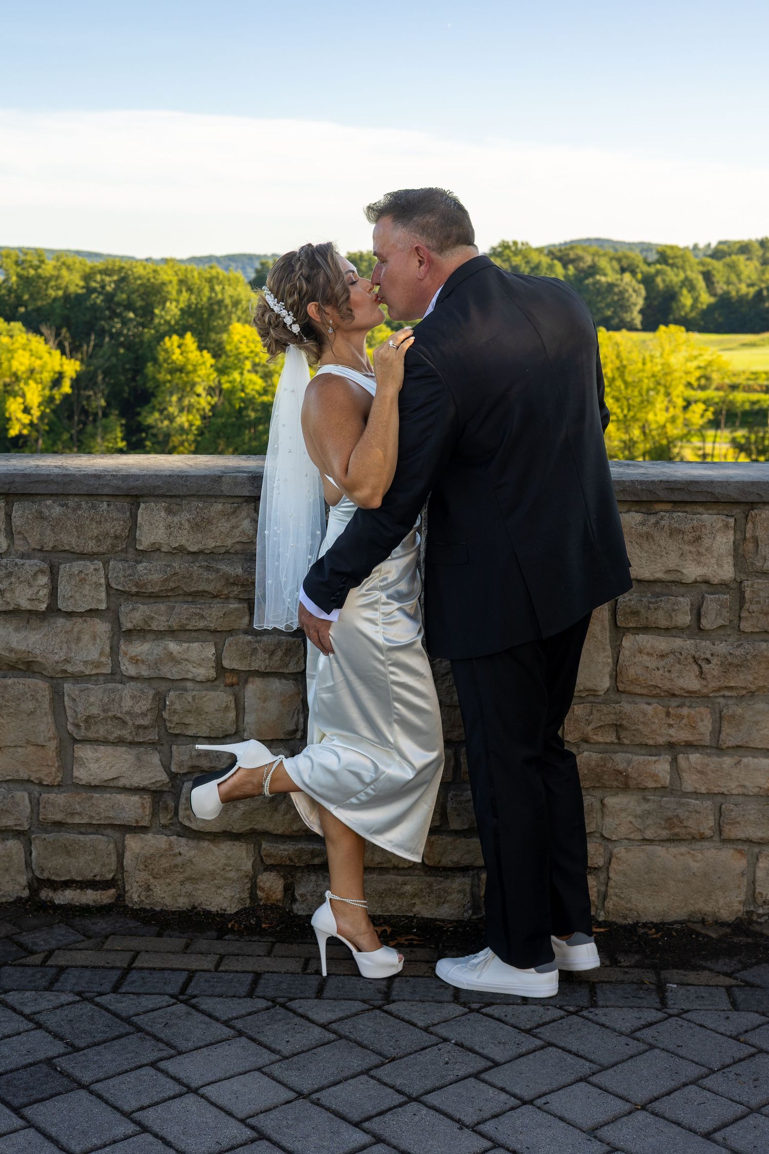 Bride and groom sharing a romantic kiss outdoors near a stone wall with lush green hills in the background.