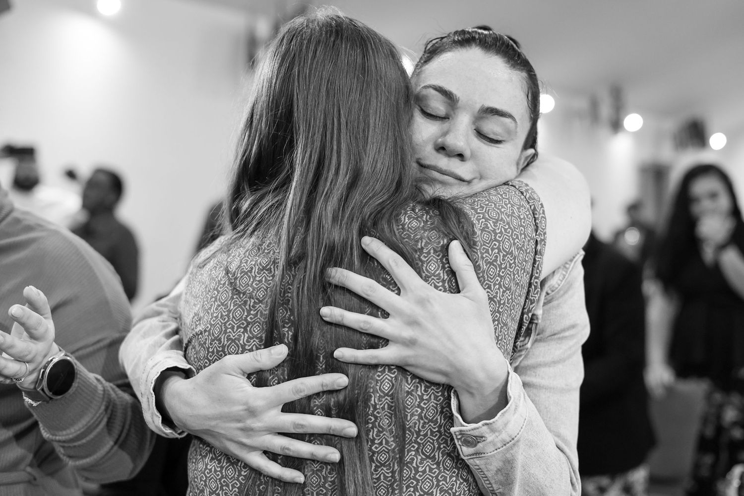 Two women share a warm embrace in a black and white candid photo at a social gathering.