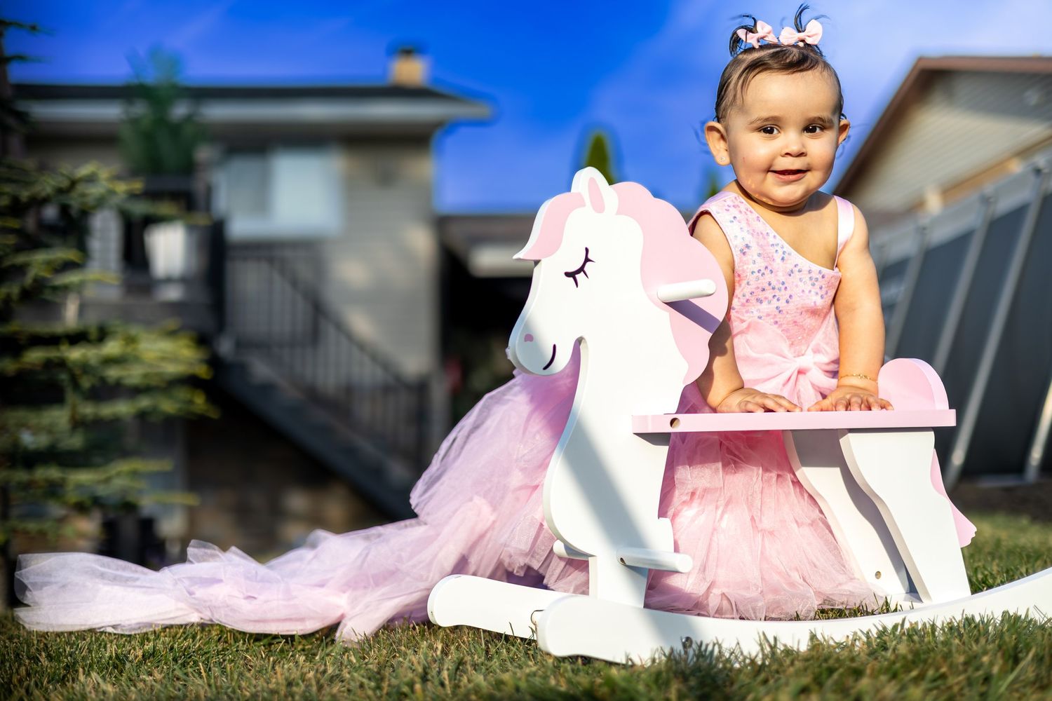Adorable baby girl in pink dress posing with white unicorn rocking horse on green lawn.