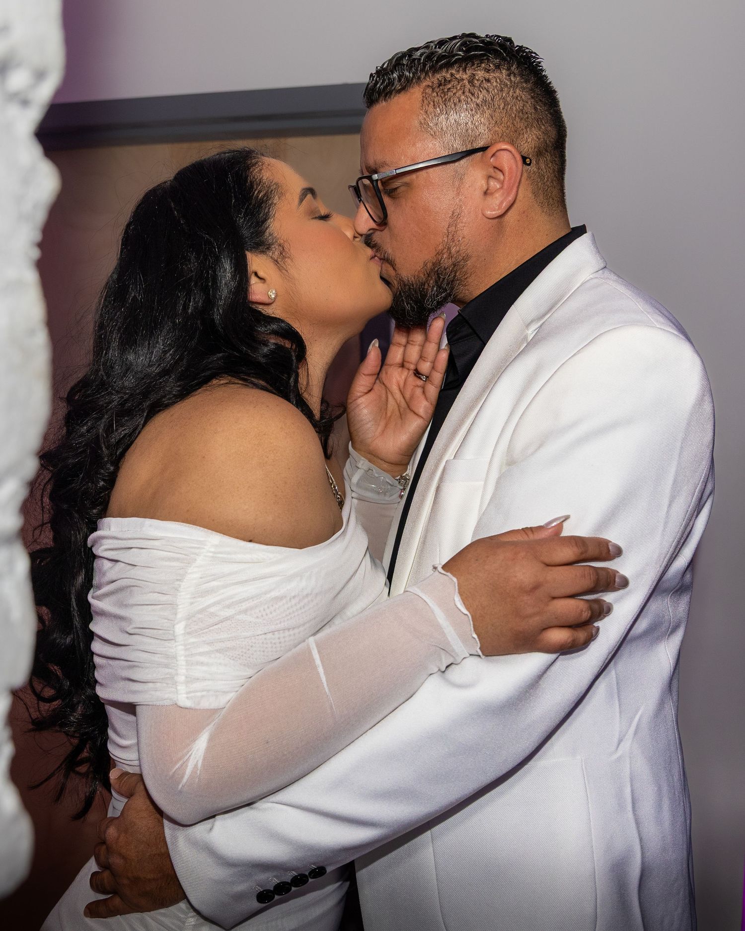 Couple sharing a romantic kiss at their wedding, both dressed in elegant white outfits, in a softly lit venue.