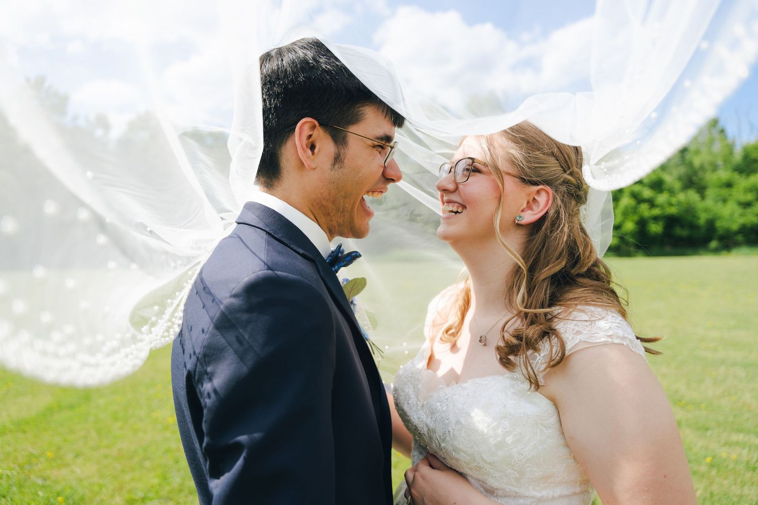 A joyful bride and groom share a romantic moment outdoors, framed by a flowing white veil on their wedding day.