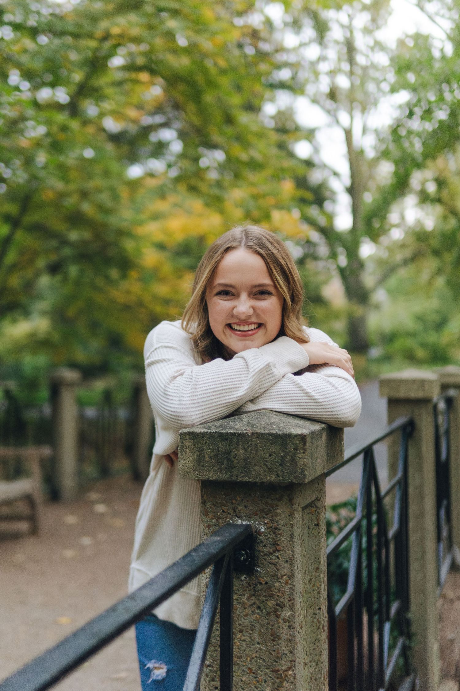 Smiling young woman leaning on stone fence post in a lush green park wearing a cream sweater and ripped jeans.