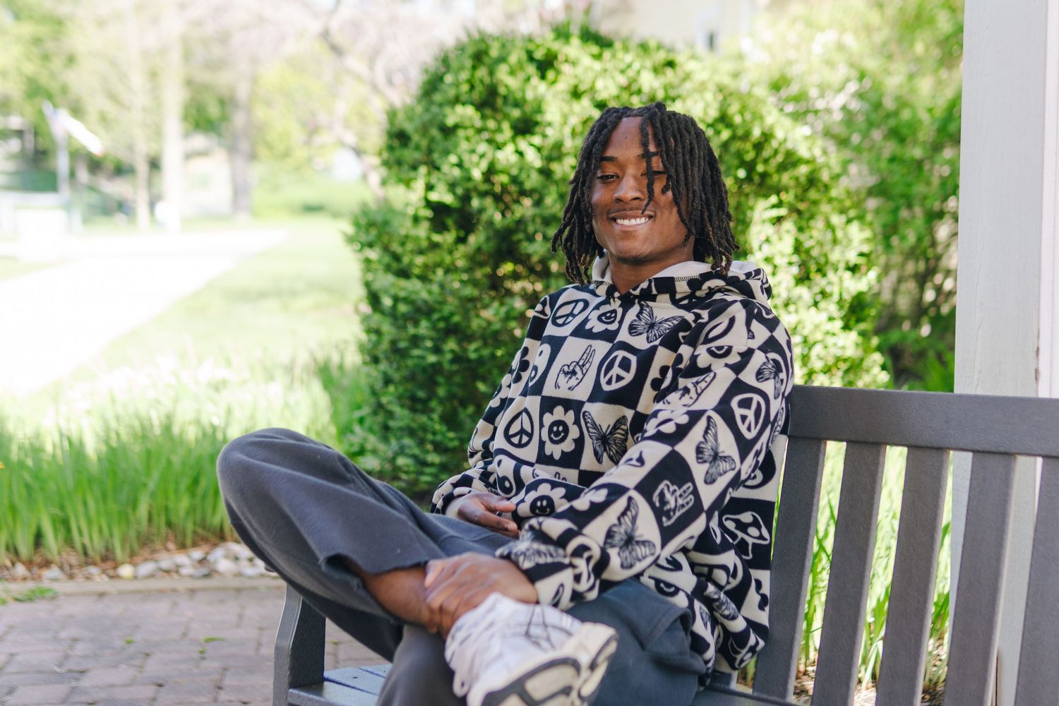 Young man smiling, sitting cross-legged on a wooden bench outdoors, wearing a patterned hoodie and gray pants.