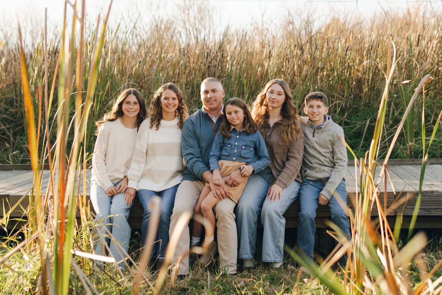 Family of six posing on a wooden dock surrounded by tall golden grasses in a warm autumn outdoor setting.