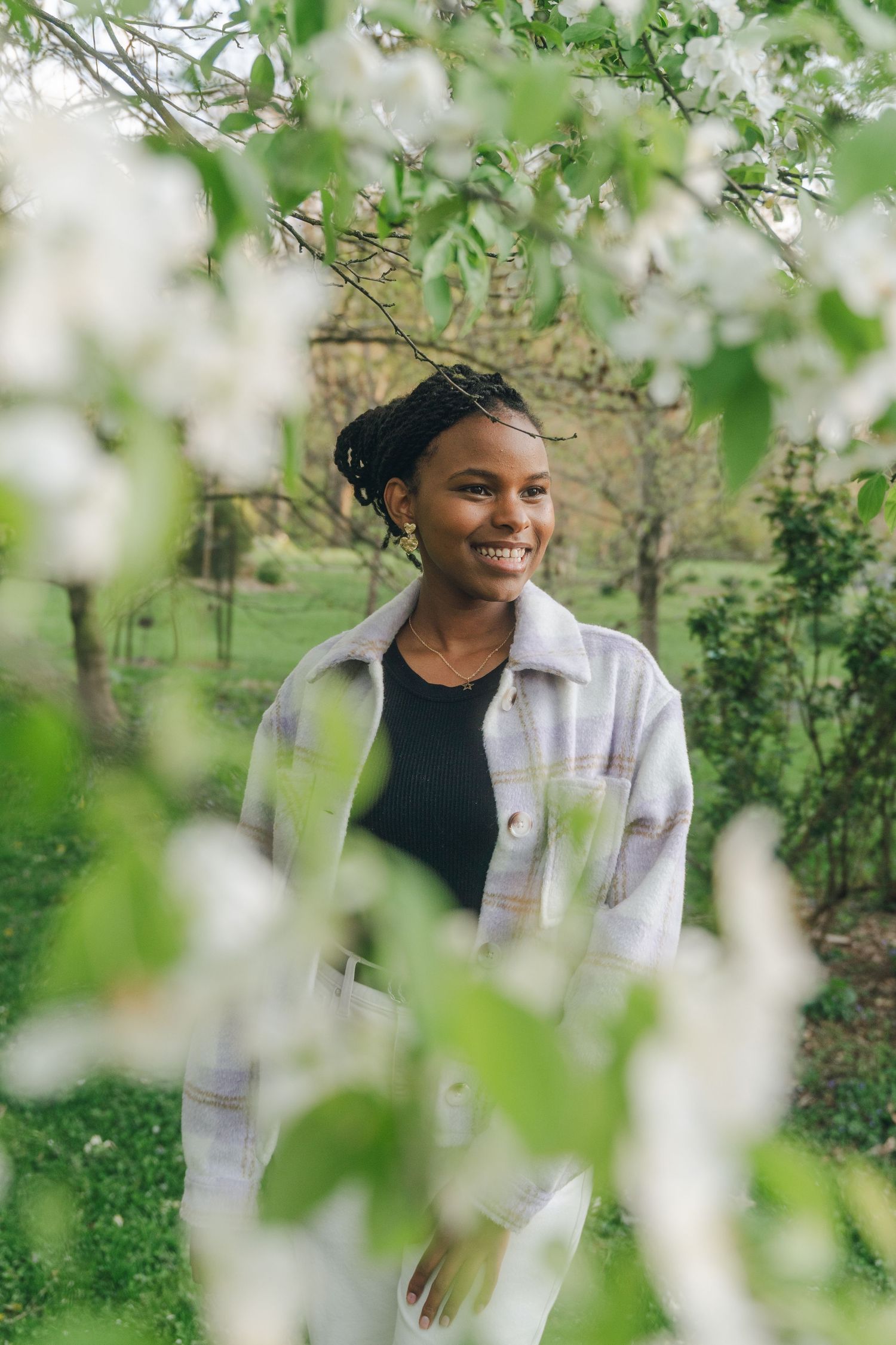Smiling young Black woman with braids standing among white blossoming trees in a green park, wearing a plaid shirt.