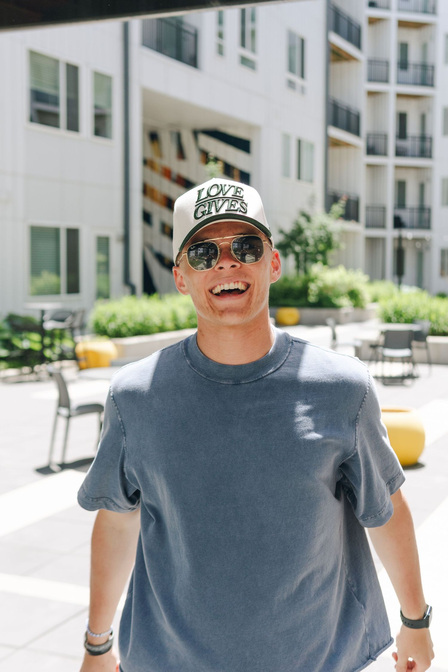 Smiling man wearing sunglasses and a Love Gives snapback cap in a gray t-shirt standing outdoors near modern apartments.