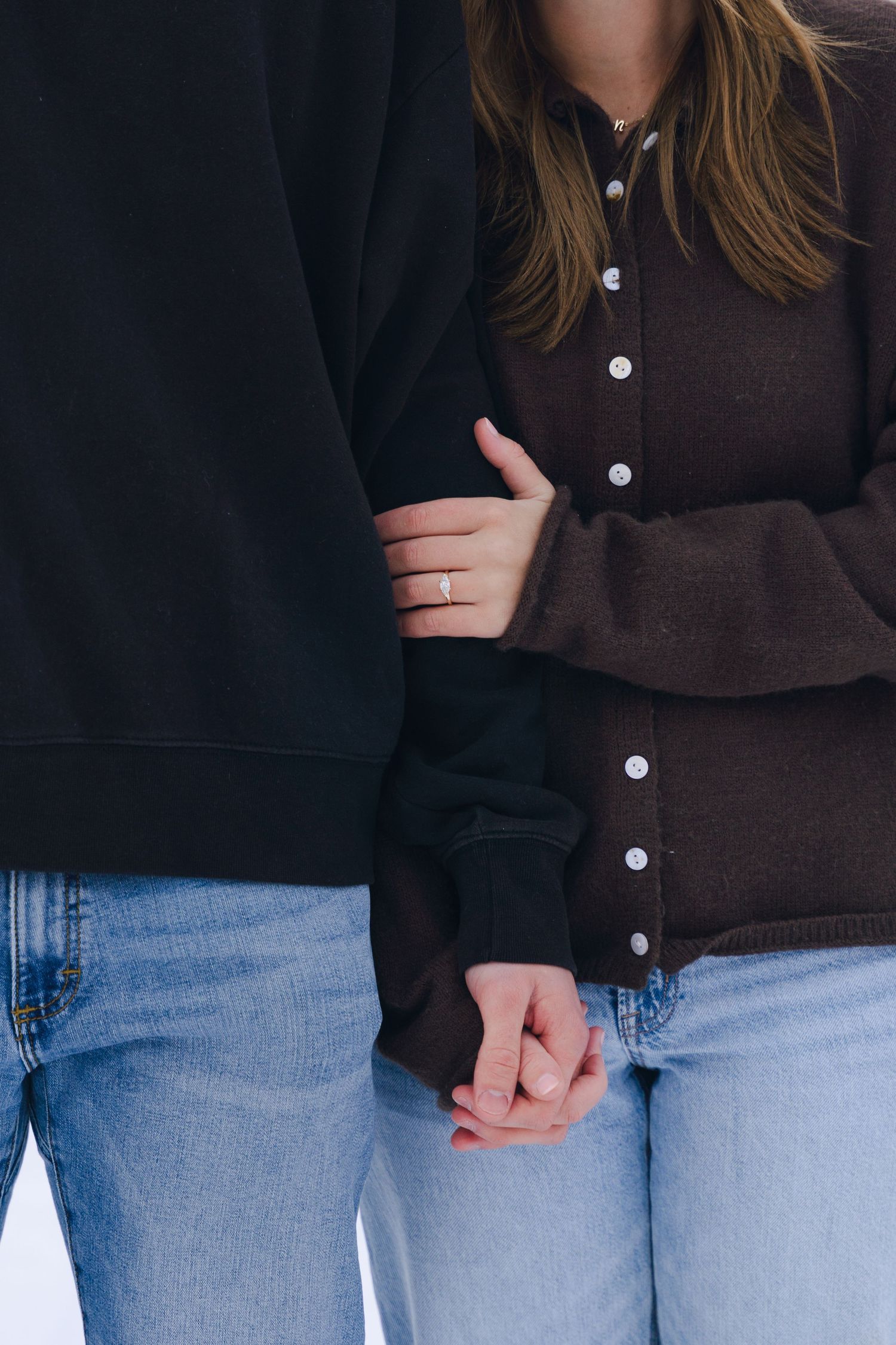 A couple holding hands and linking arms, wearing jeans and dark sweaters, with a ring visible on the woman's finger. proposal photoshoot