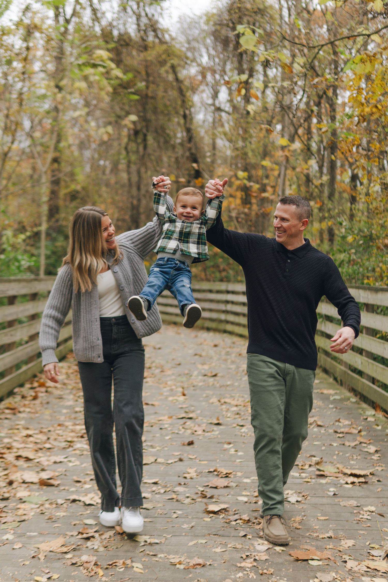 A happy family walks on a wooden bridge in autumn, parents swinging their young child by the hands.