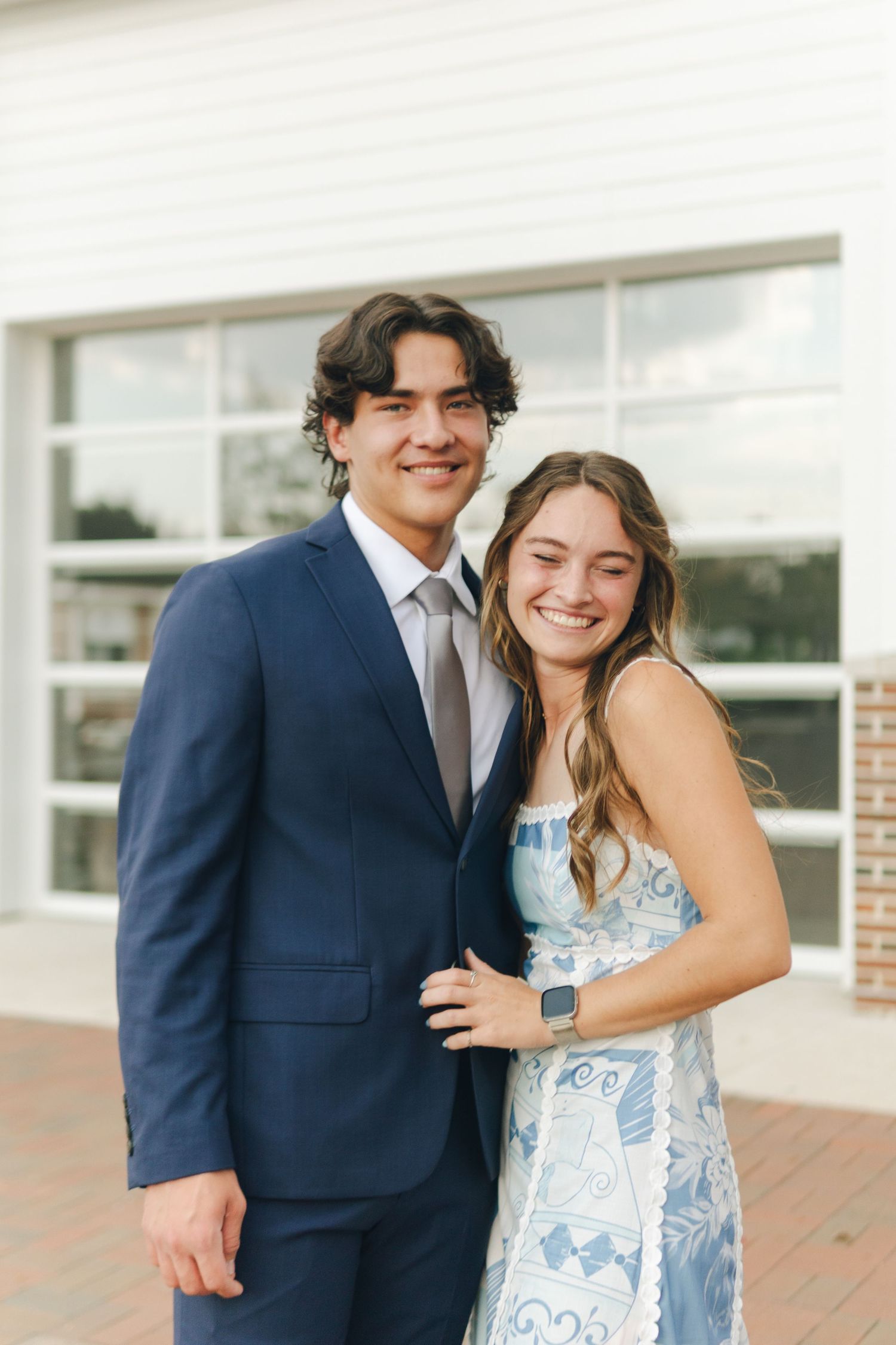 Smiling young couple posing outdoors, man in navy suit and woman in blue floral dress.