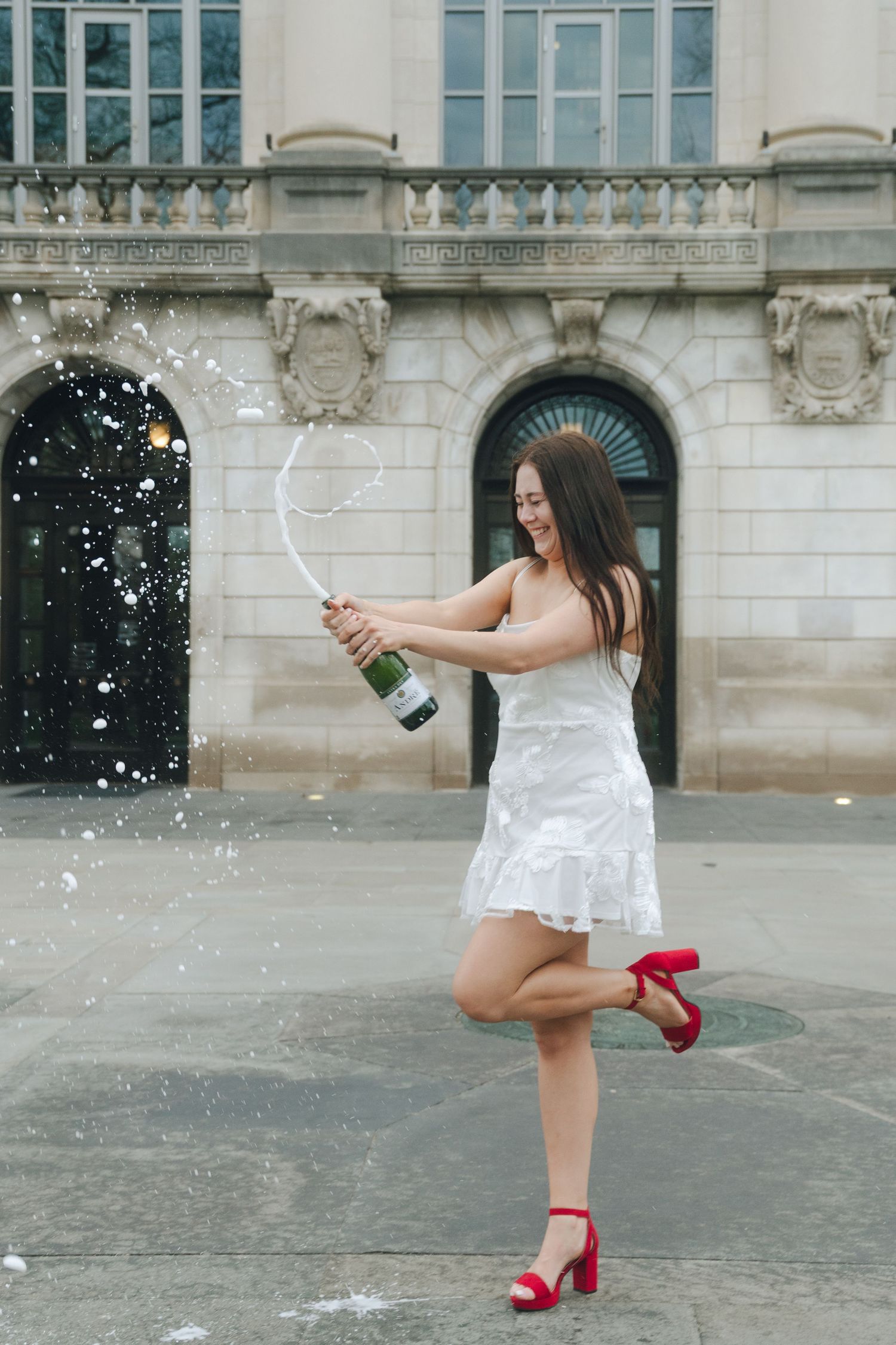 Young woman in white dress and red heels celebrating outdoors, popping champagne bottle in front of elegant building.
