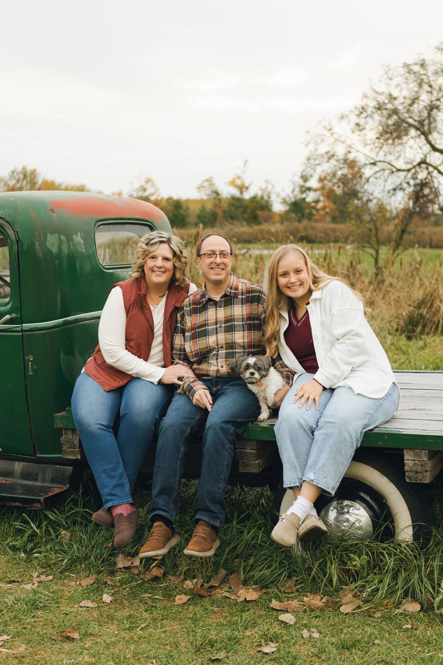 Family of three smiling on vintage green truck tailgate in autumn field, holding small dog, wearing casual fall outfits.
