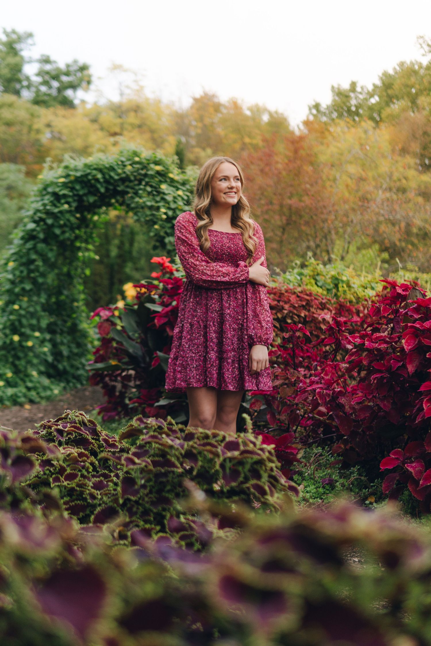 Young woman in floral burgundy dress standing in a lush garden with red foliage and green ivy arch.