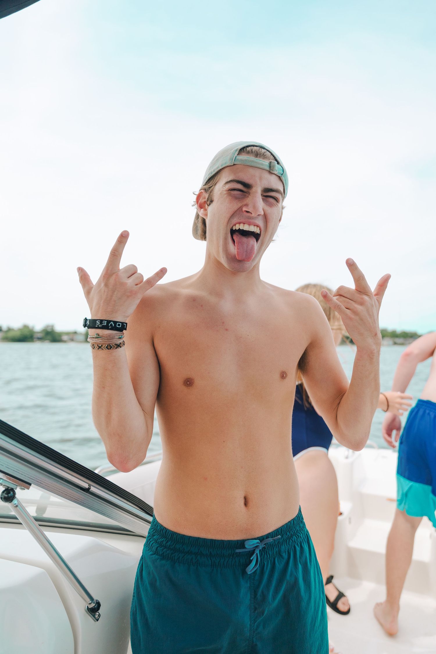 Shirtless young man on a boat making rock on hand gestures with tongue out, wearing teal swim trunks and backwards cap.