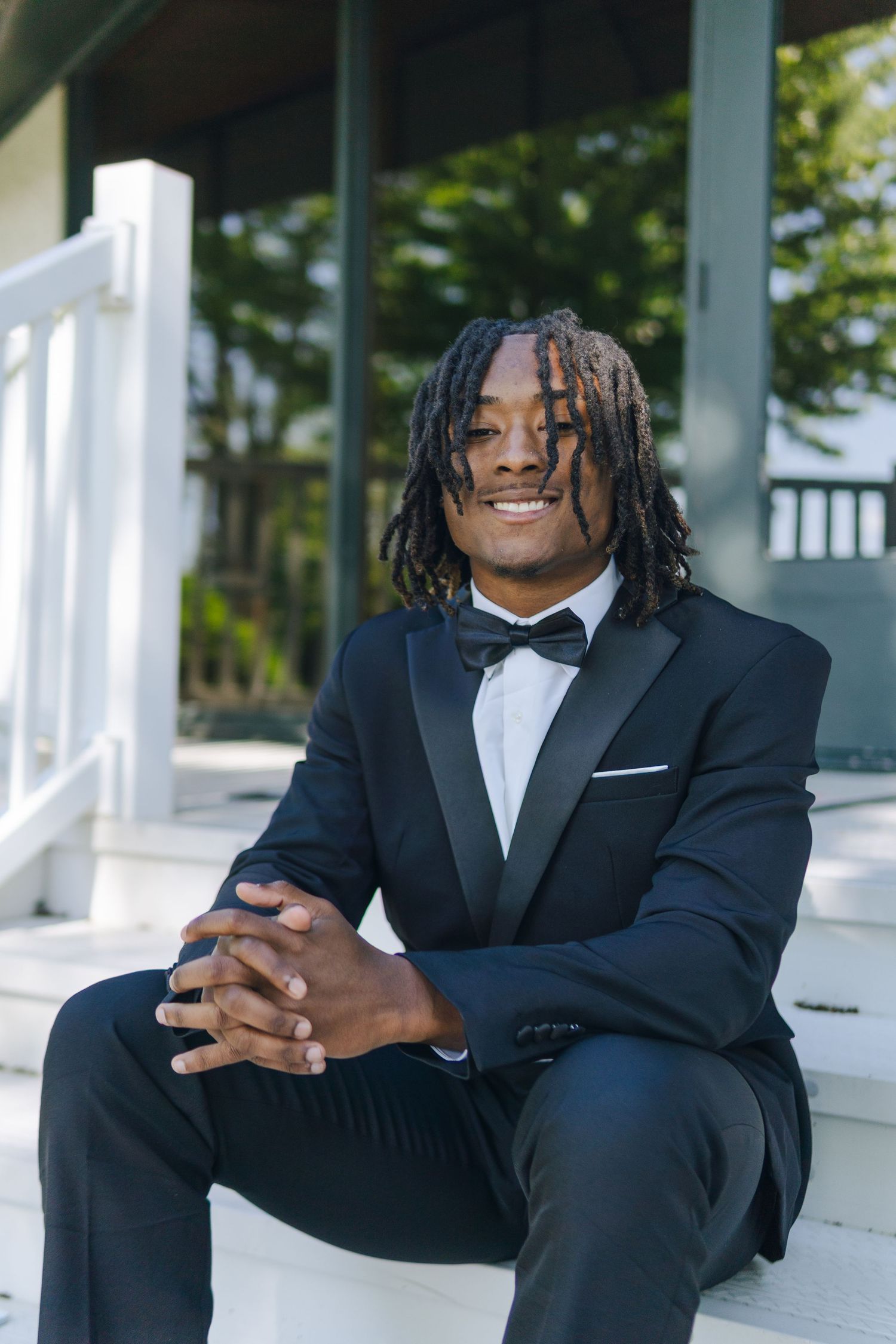 Young man in black tuxedo with bow tie sitting on white steps, smiling confidently with dreadlocks. Getting Senior photos taken