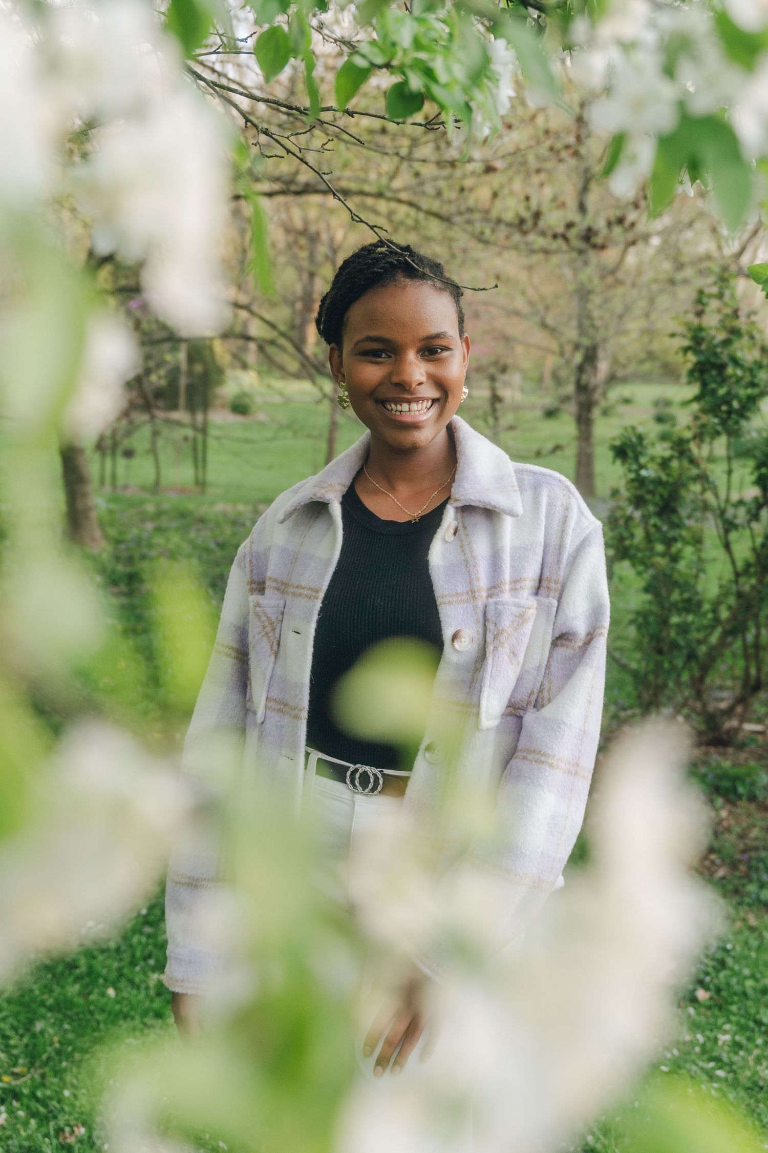 Young Black woman smiling in a blooming garden, wearing a plaid jacket over a black top, surrounded by white spring flowers.