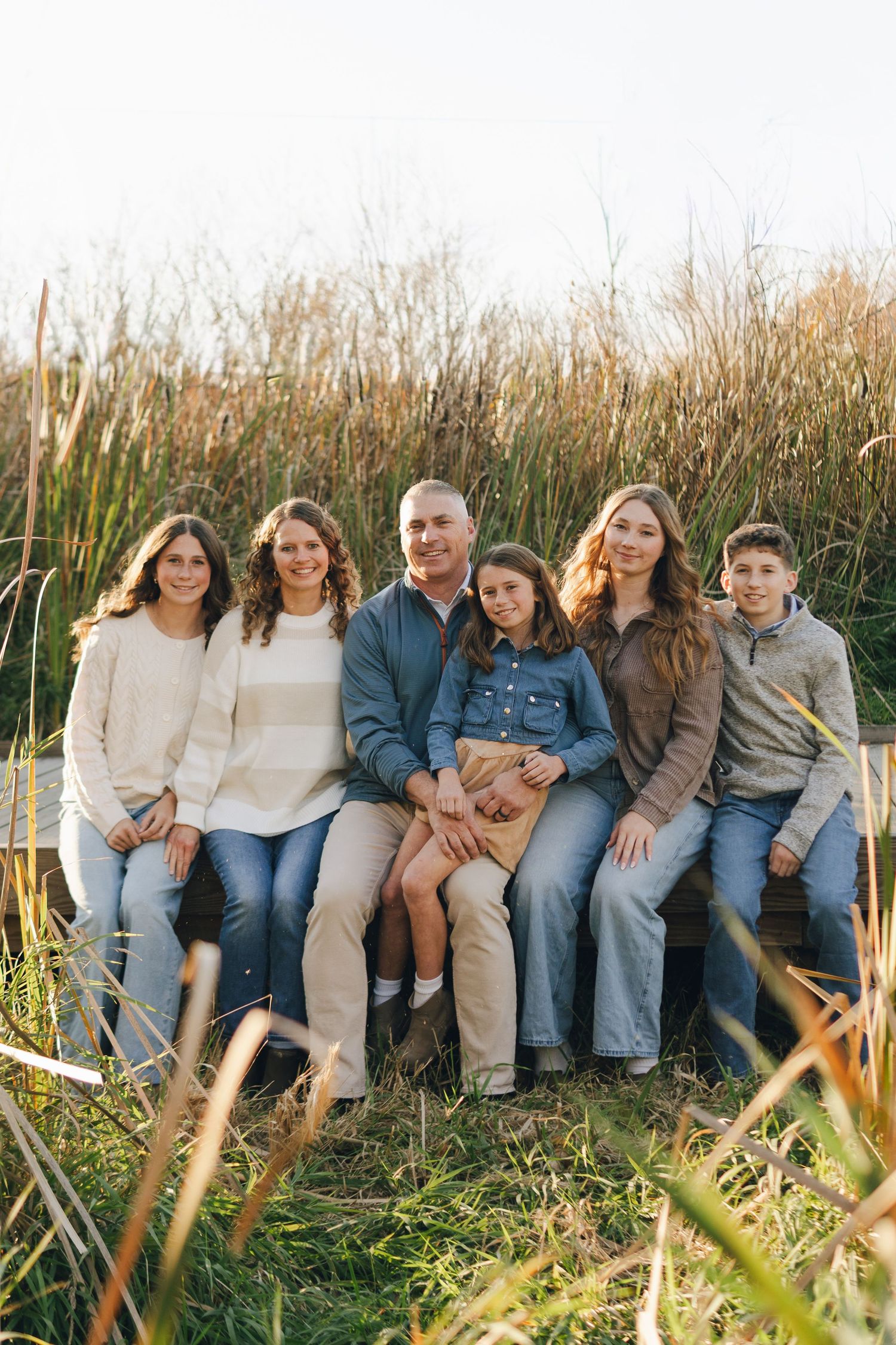 Family of six posing outdoors on a bench surrounded by tall golden grass in a warm autumn field setting.