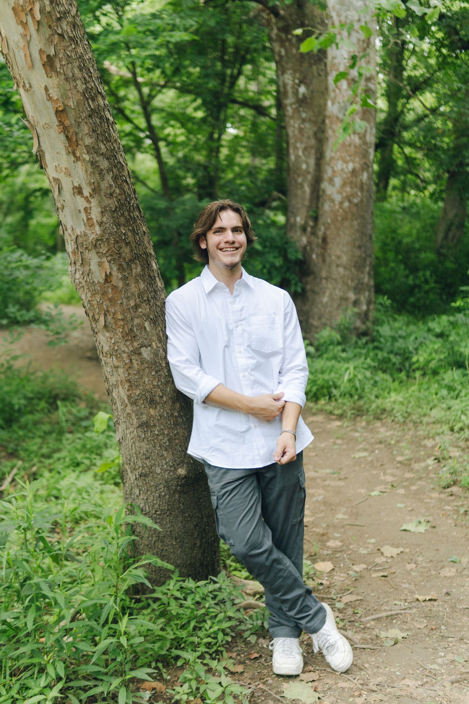 Young man in white shirt leaning against tree on forest path, smiling outdoors.