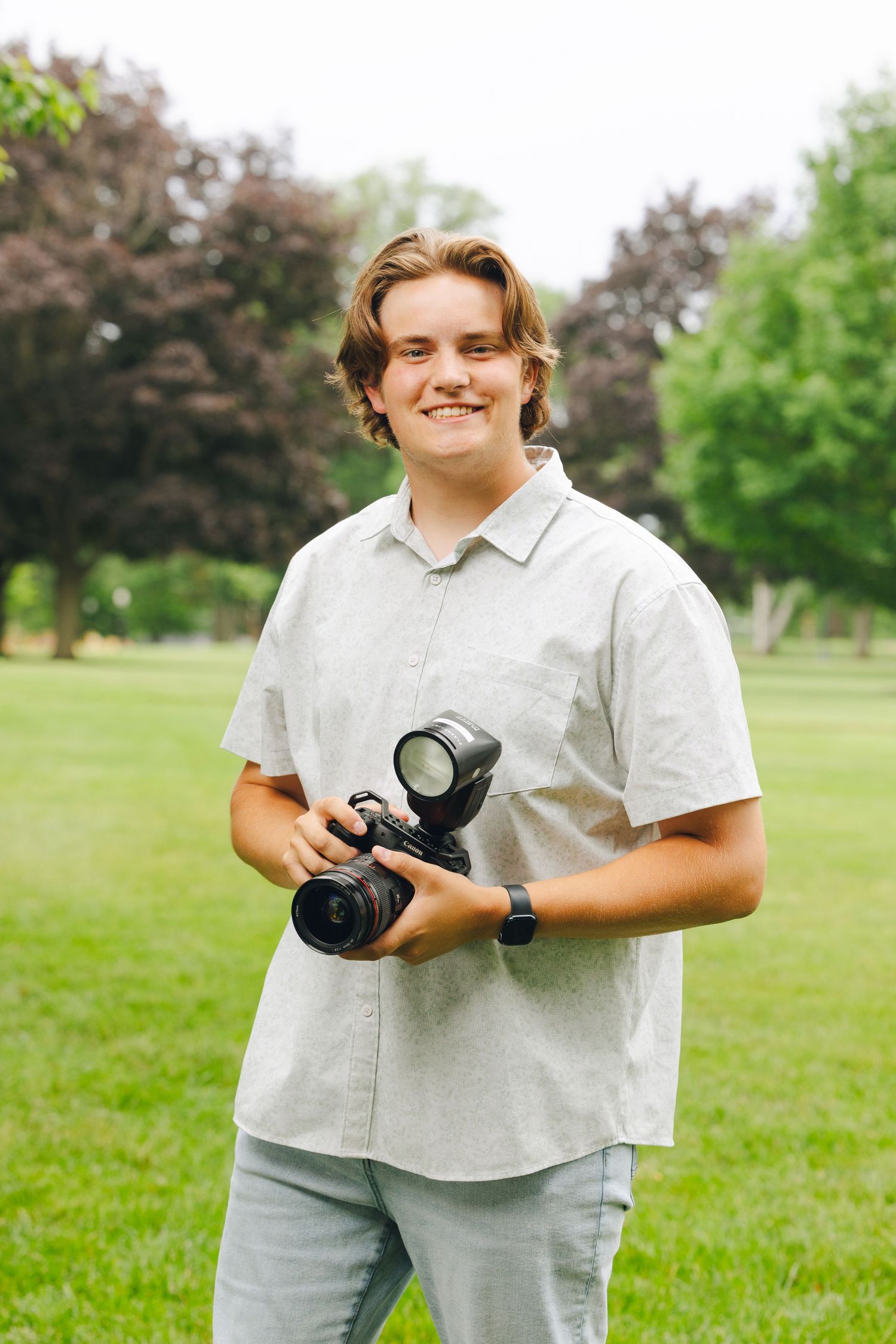 Smiling young man holding a DSLR camera with flash outdoors in a green park wearing a light gray shirt.