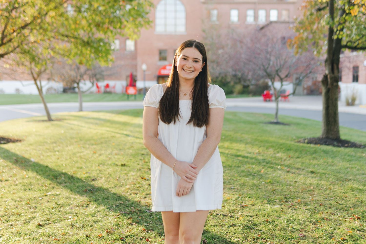 Young woman in white dress smiling outdoors on college campus green lawn with brick building background.