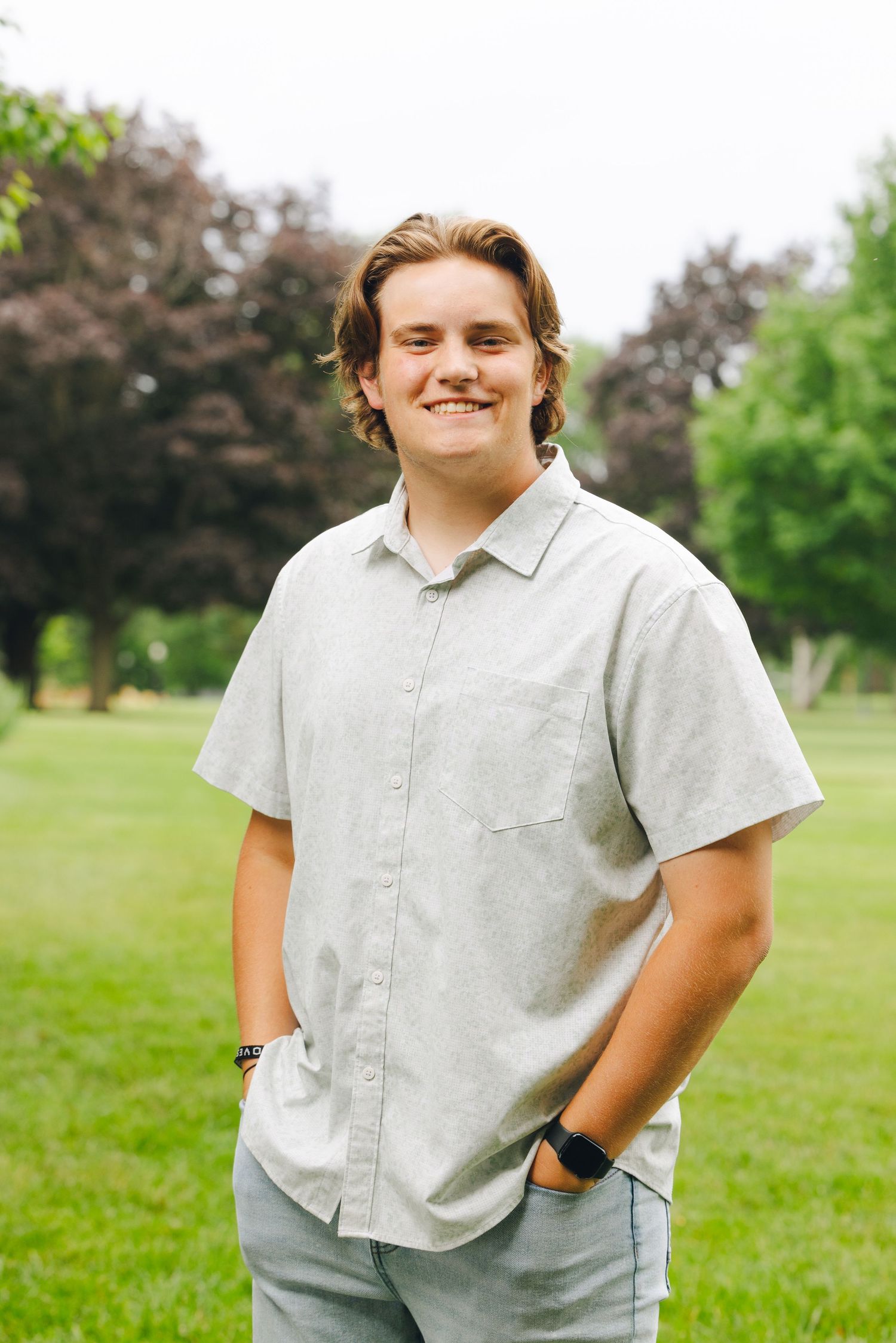 Smiling young man in a light gray button-up shirt standing in a green park with trees in the background.