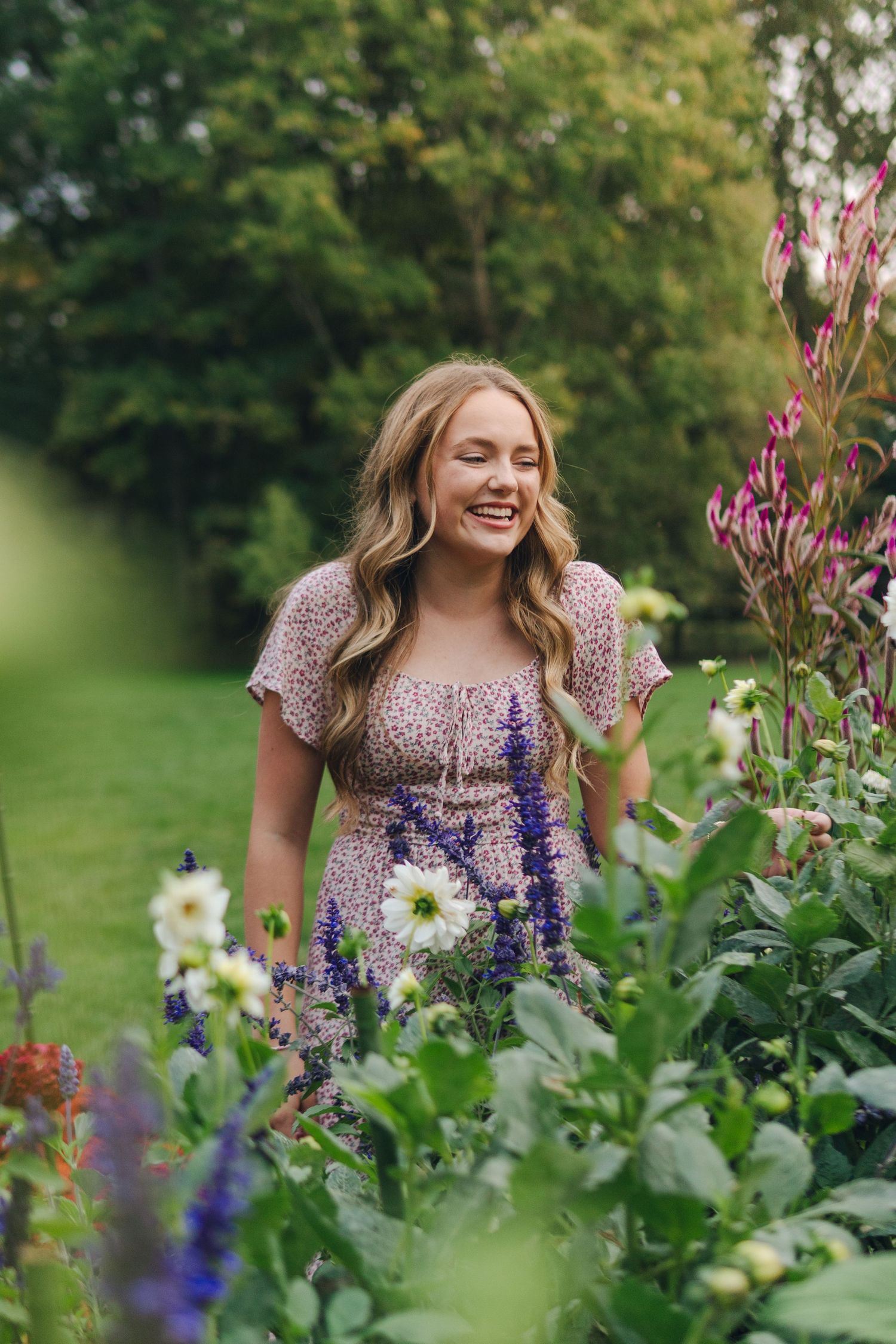Smiling blonde woman in floral dress standing among colorful garden flowers with lush green trees in background.