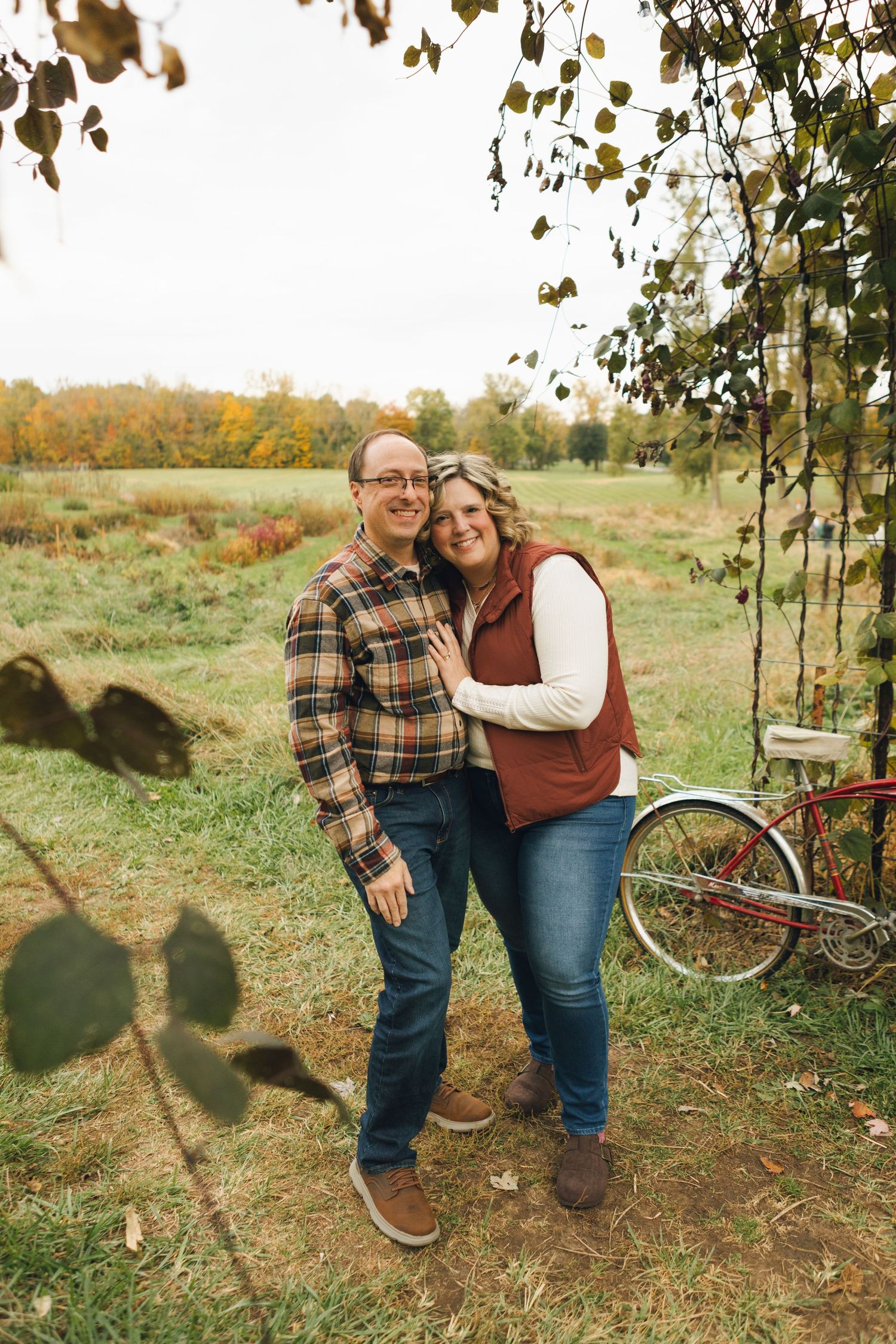 Couple embracing outdoors in autumn, wearing flannel and vest, with a red bicycle and fall foliage in the background.