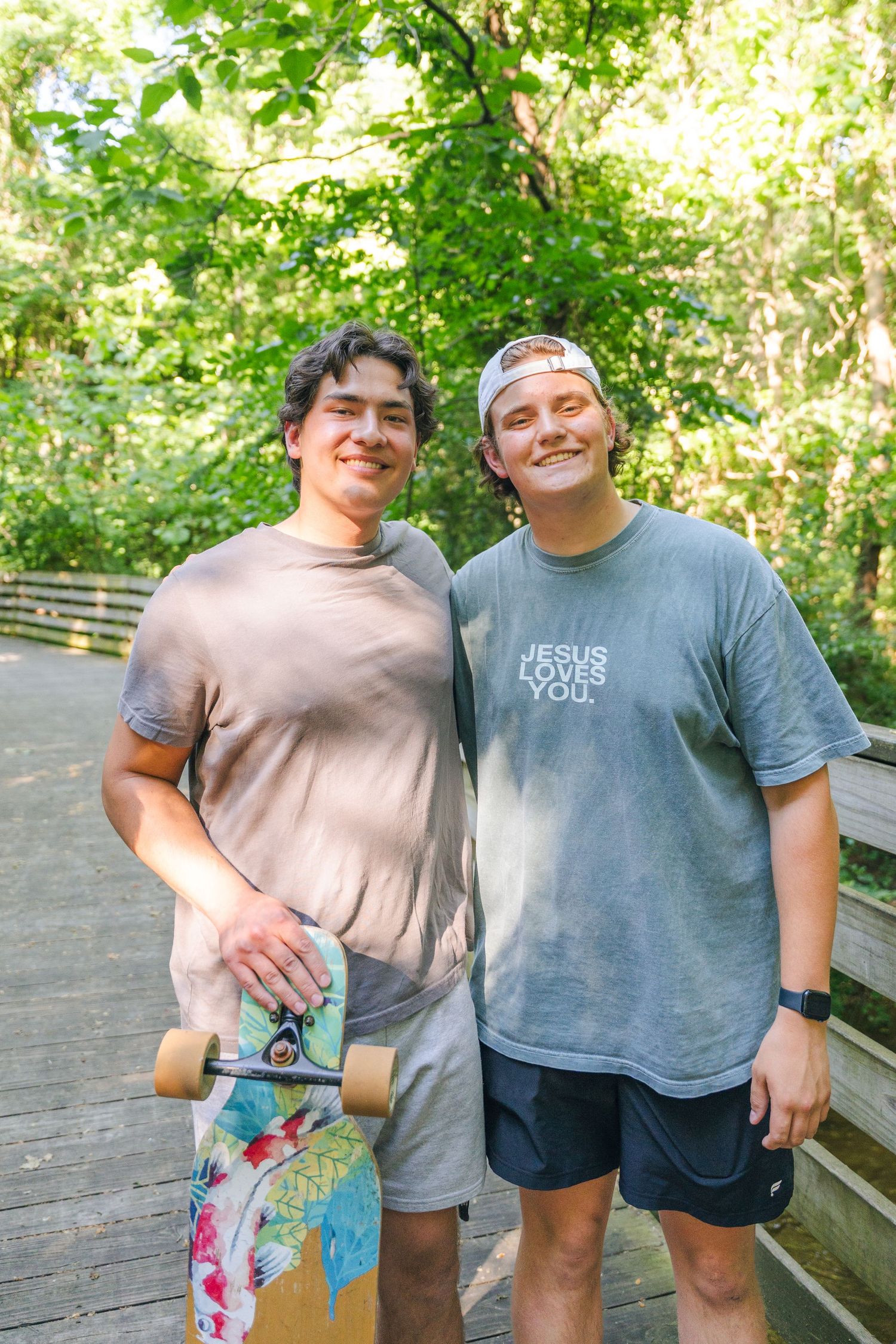 Two smiling young men pose on a wooden bridge outdoors, one holding a colorful skateboard, wearing casual t-shirts.