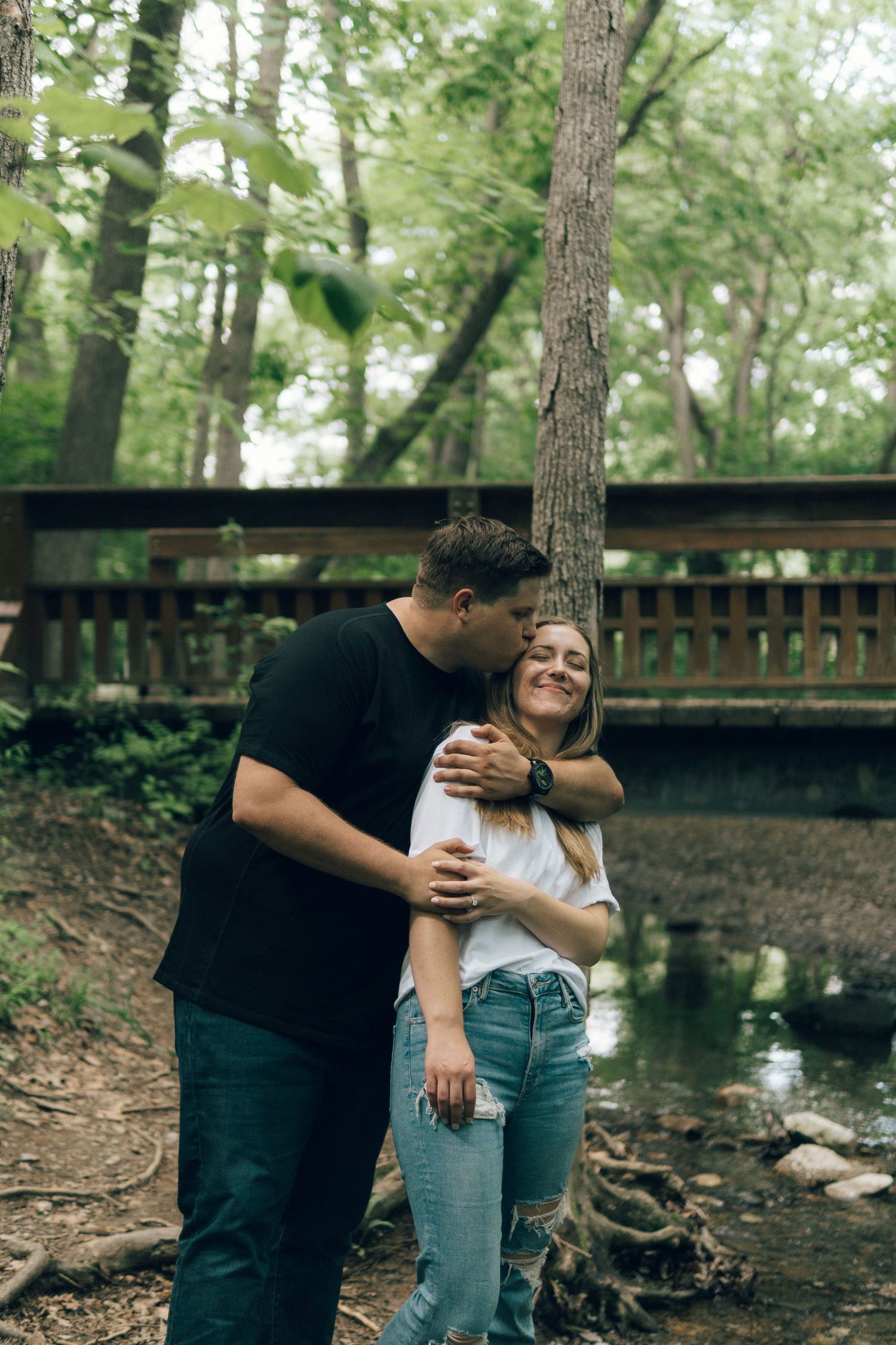 A couple embraces near a wooden bridge in a lush forest, the man kissing the smiling woman on the head.