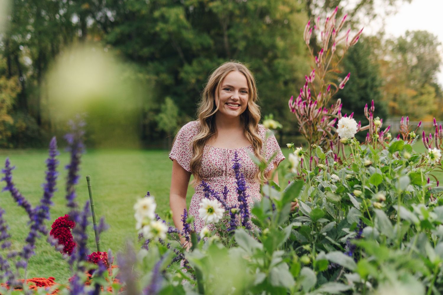 Smiling woman in floral dress standing among colorful garden flowers with lush green trees in background.