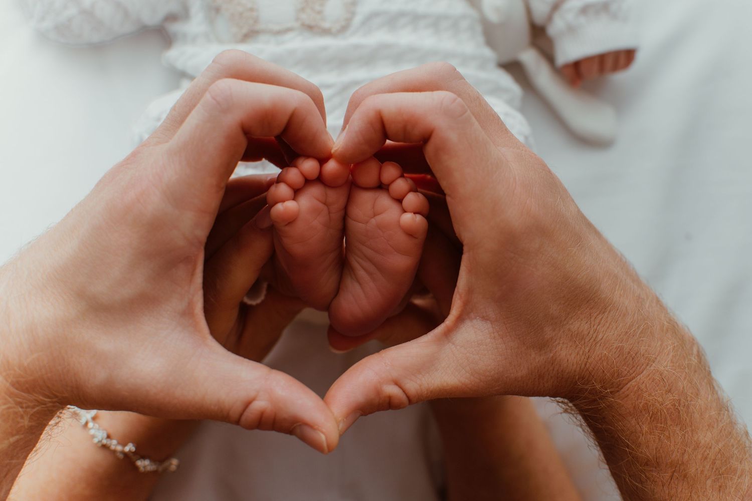Two parents form a heart shape with their hands around their newborn baby's tiny feet in a tender moment.