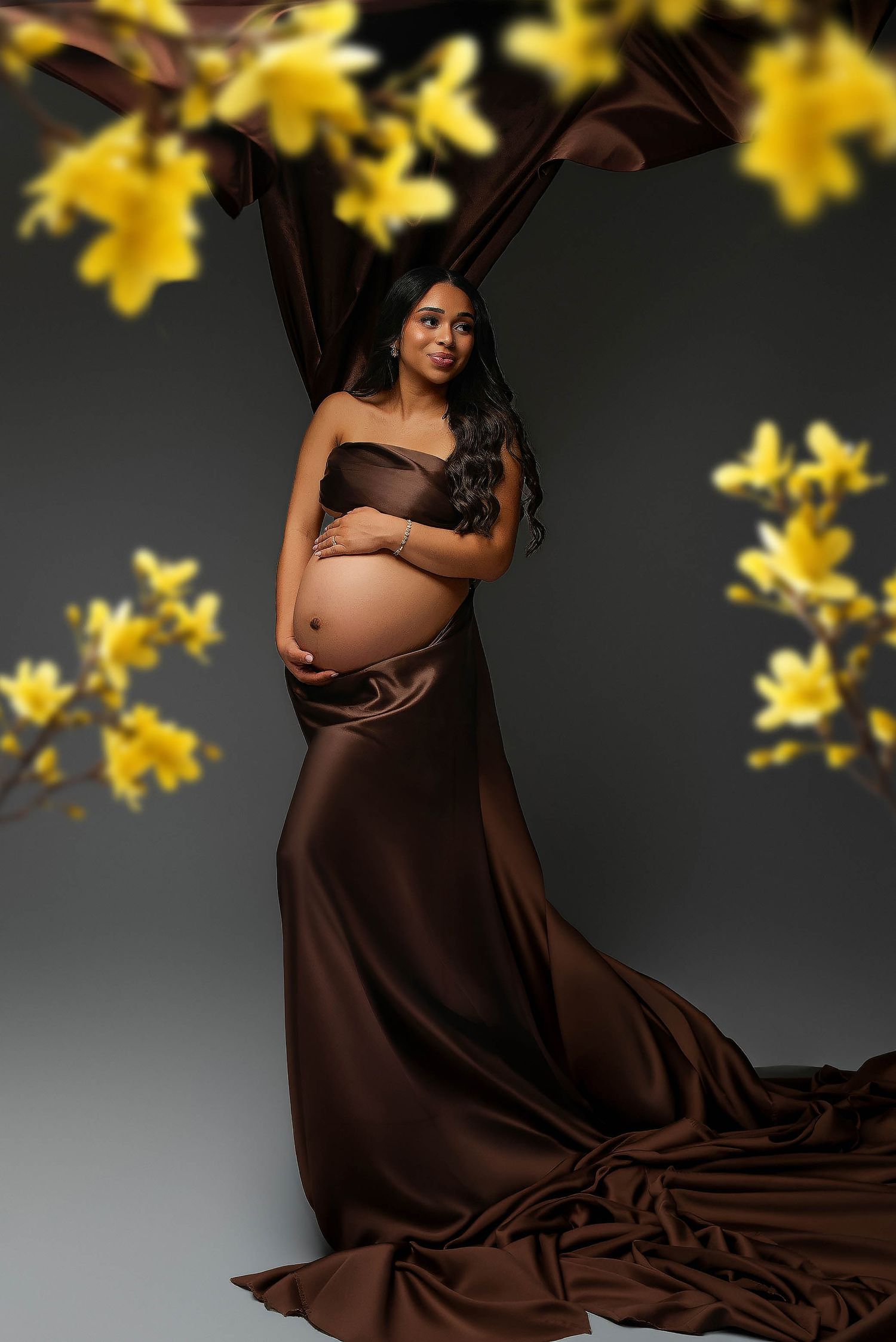 Pregnant woman in elegant brown satin gown poses gracefully among yellow forsythia branches in a studio maternity portrait.