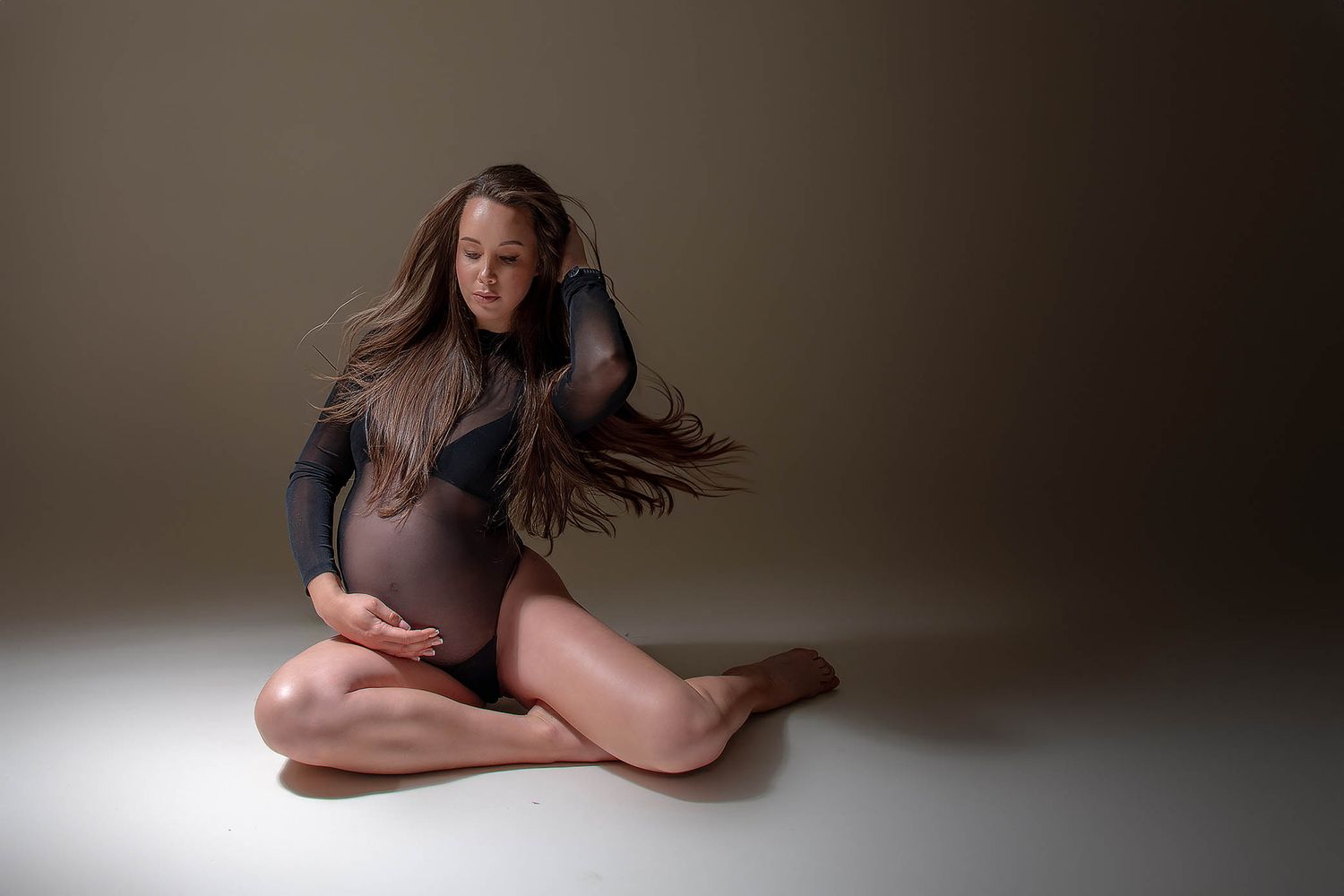 Pregnant woman in sheer bodysuit sitting gracefully on studio floor, flowing hair, cradling baby bump in artistic maternity photo.