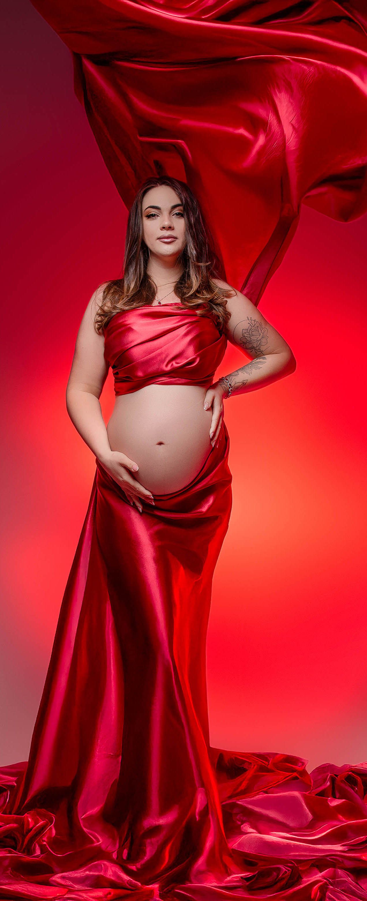 Pregnant woman in red satin two-piece maternity gown with flowing fabric on red background.
