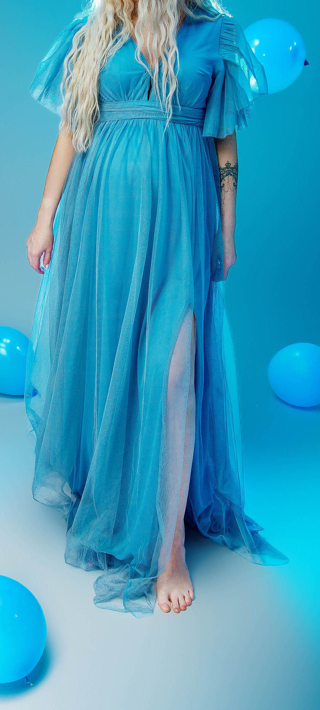 Woman in flowing blue tulle gown with flutter sleeves and front slit, surrounded by blue balloons on blue background.
