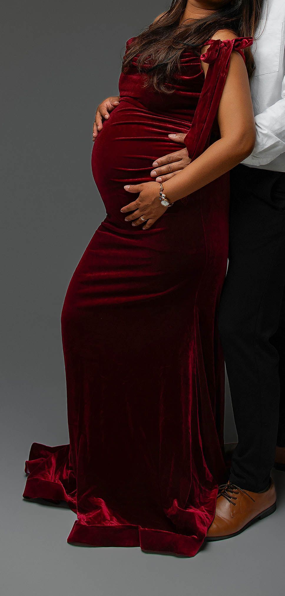 Pregnant woman in red velvet maternity gown with partner embracing her from behind in studio.