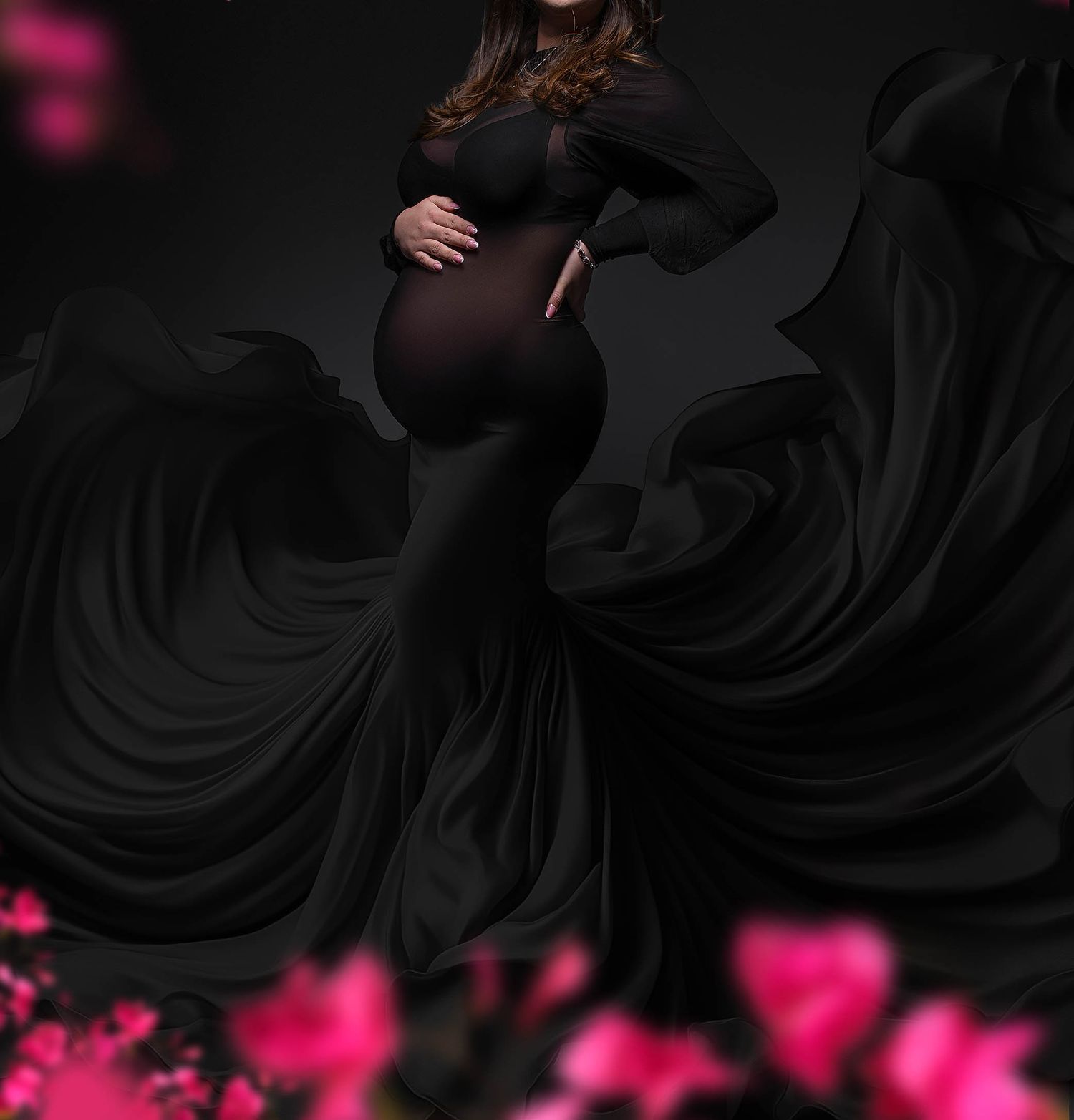 Pregnant woman in elegant black maternity gown with flowing fabric, posing against dark background with pink flowers.