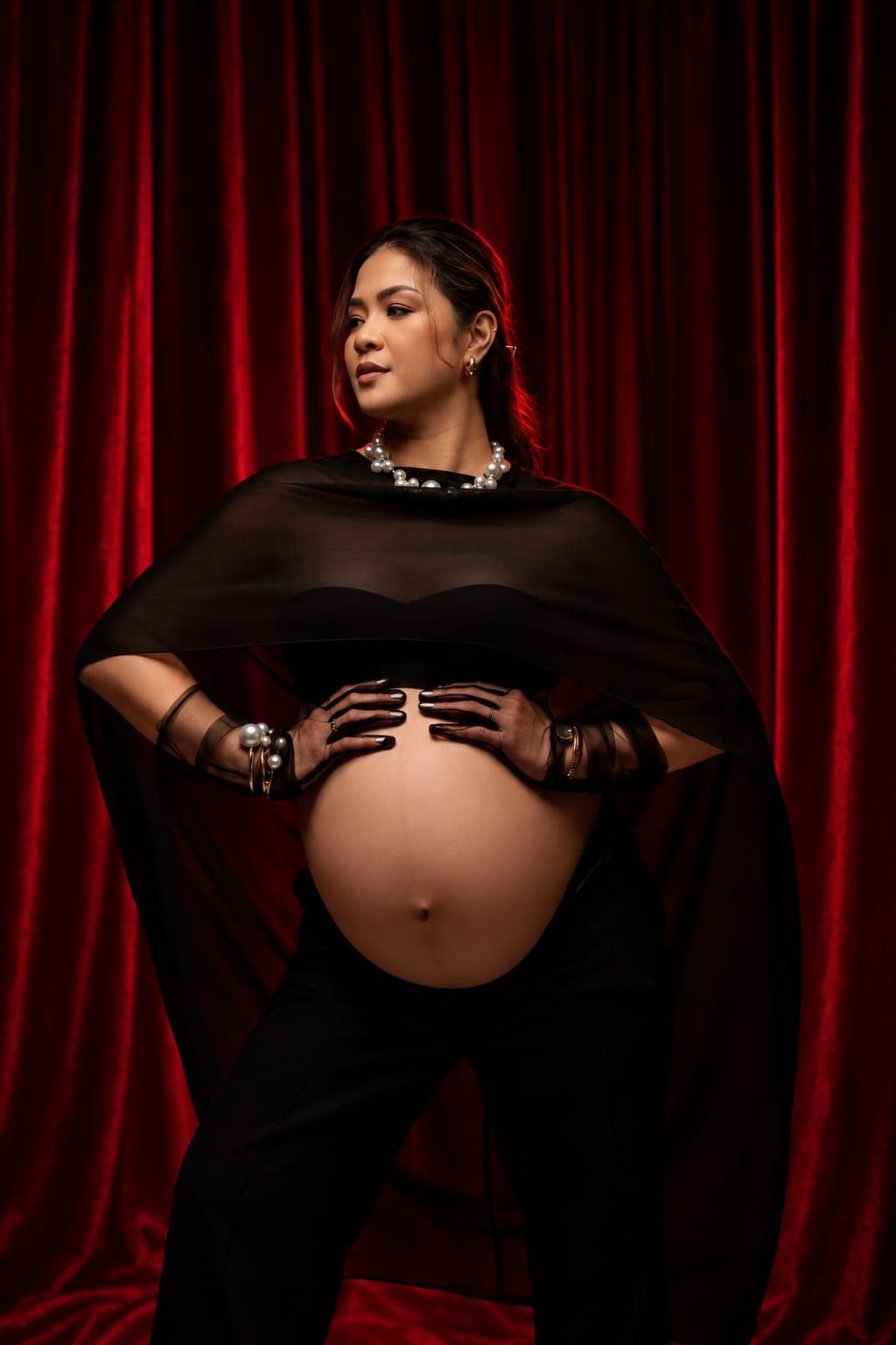 Pregnant woman in black cape and gloves posing against red velvet curtains, showcasing baby bump in elegant maternity shoot.