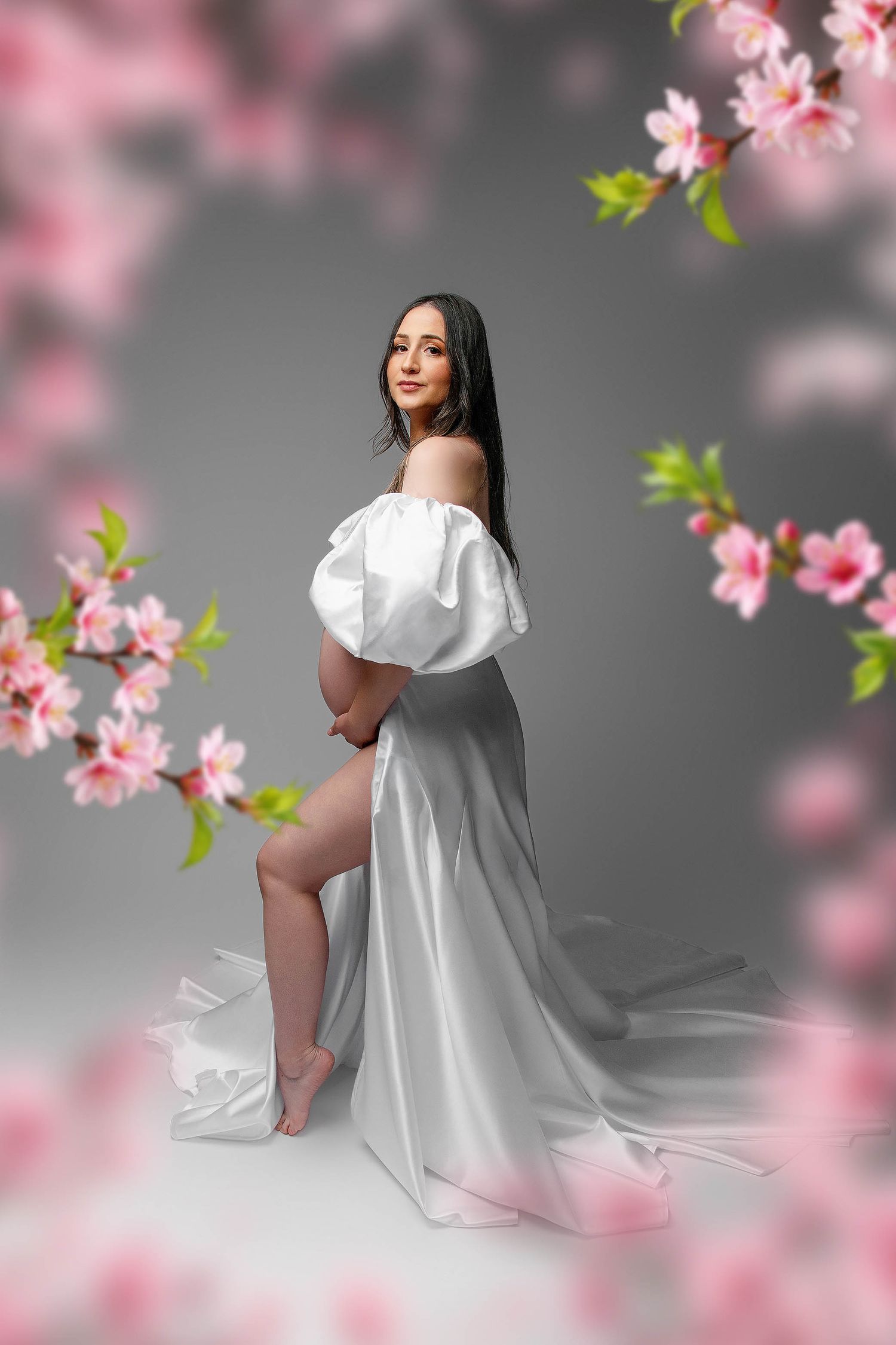 Pregnant woman in white off-shoulder gown poses gracefully among pink cherry blossoms on gray background.
