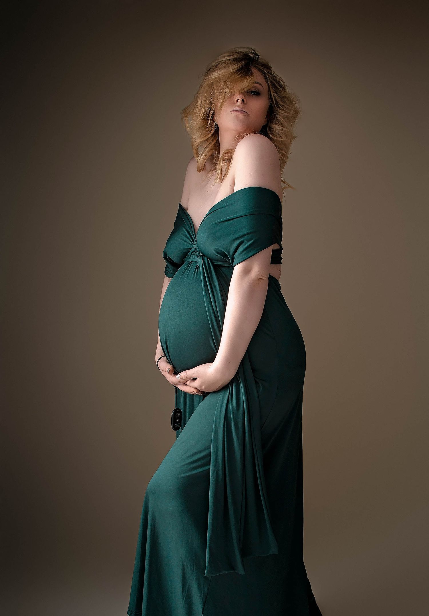 Pregnant woman in elegant off-shoulder emerald green maternity gown cradling her baby bump during a studio photoshoot.
