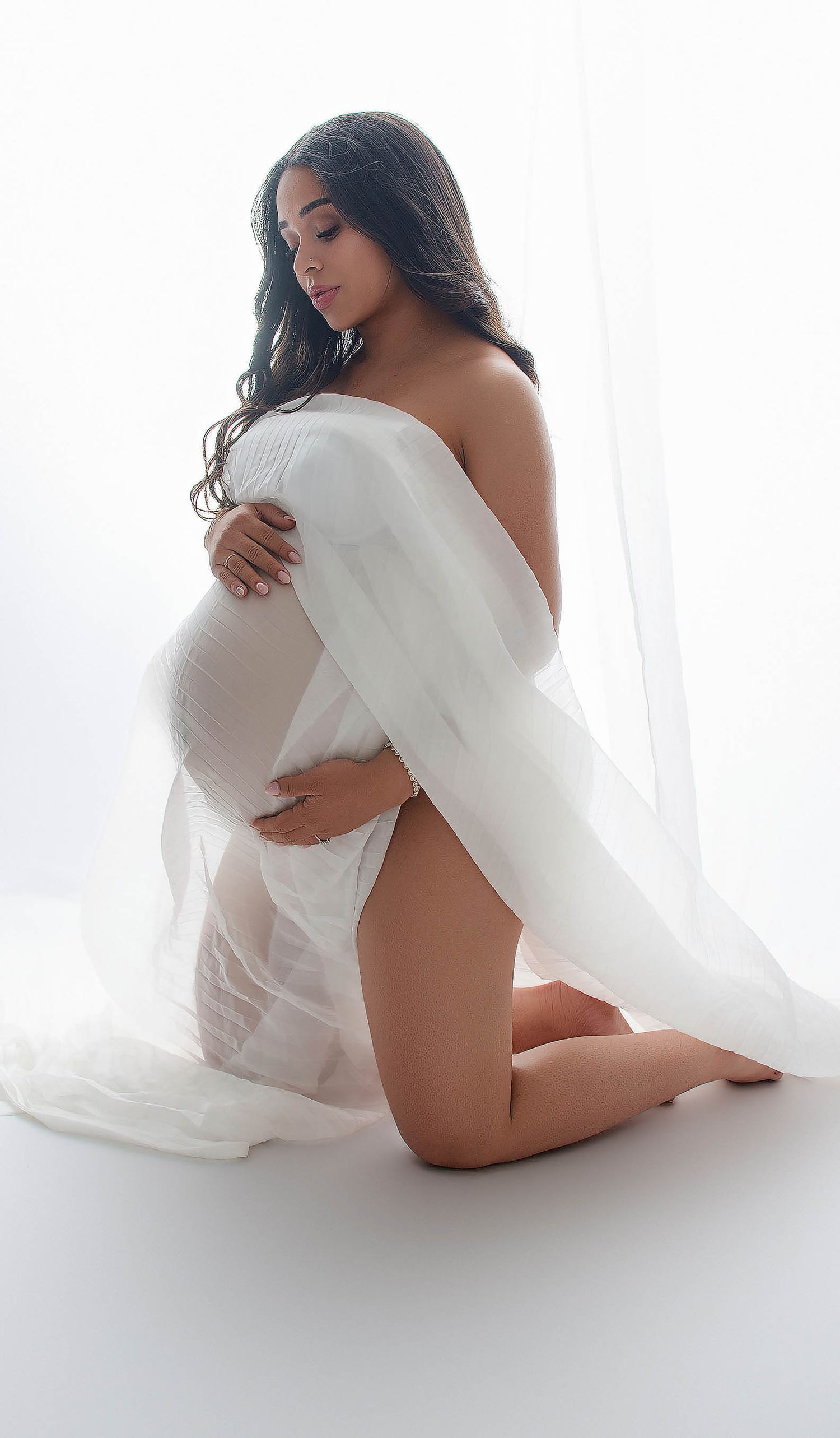 Pregnant woman kneeling gracefully draped in sheer white fabric, cradling her baby bump in a bright studio maternity photo.