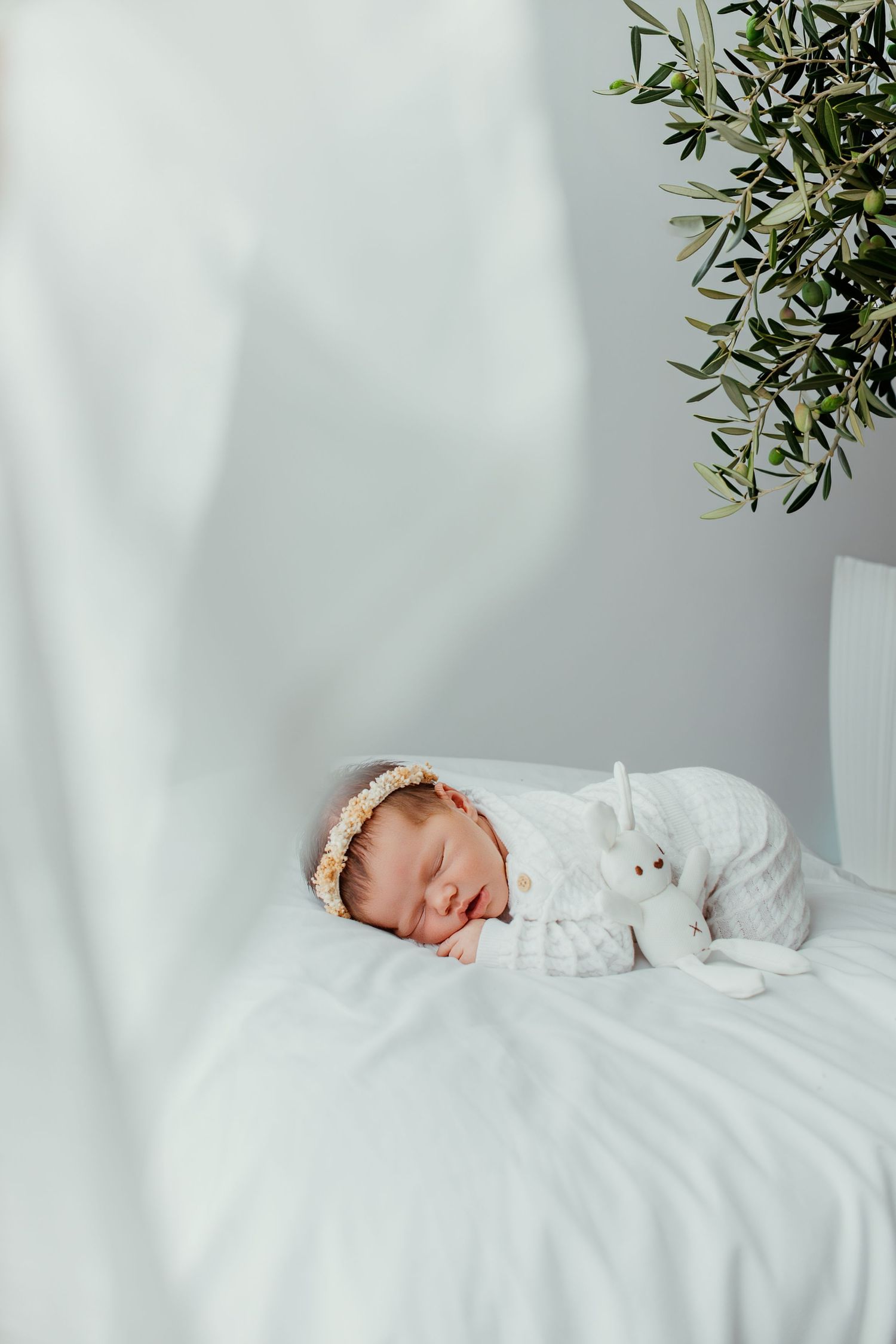 Sleeping newborn baby in white knit outfit with floral headband cuddling a bunny stuffed animal on white bedding.