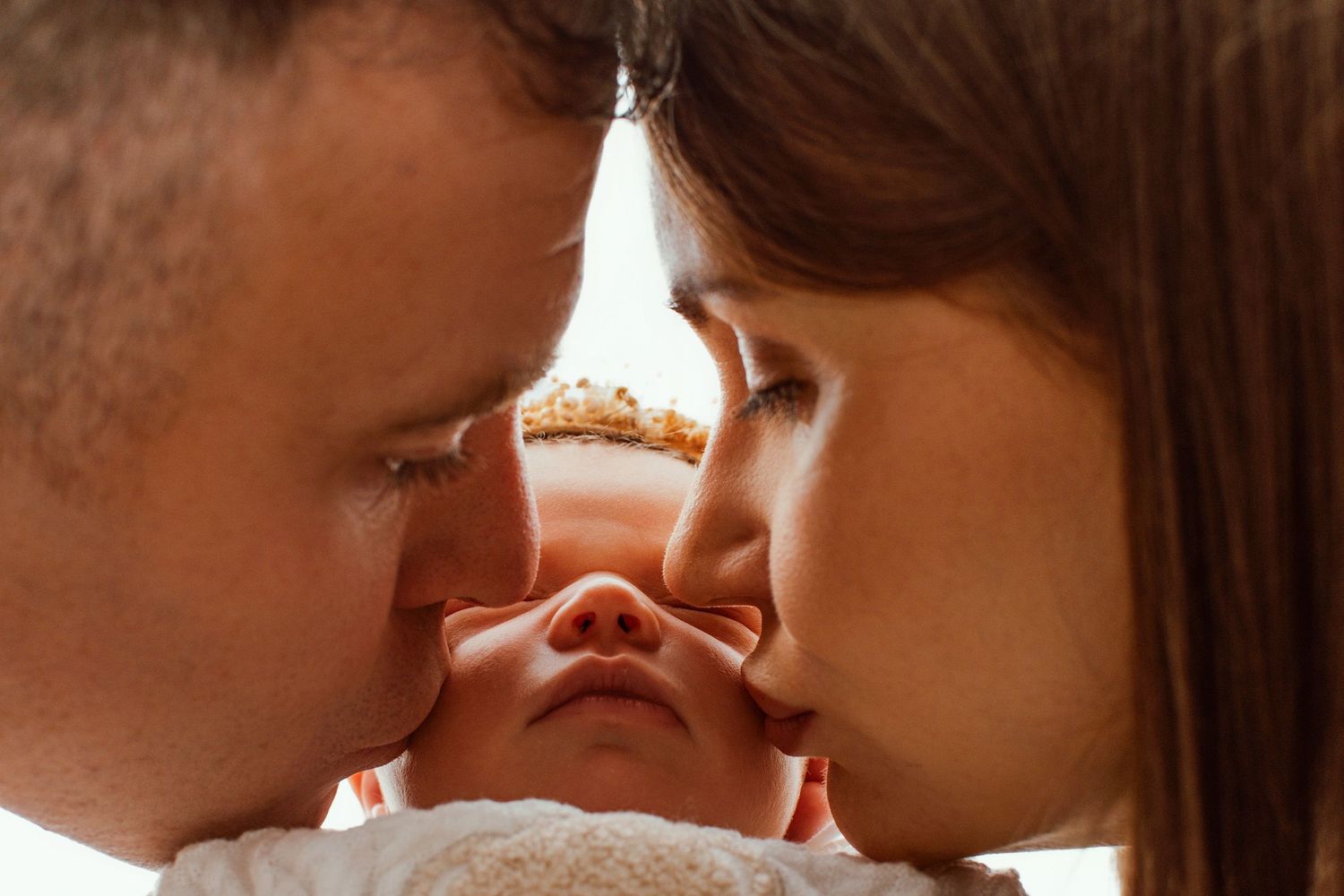 Parents kissing their newborn baby on the nose in a tender close-up family portrait.