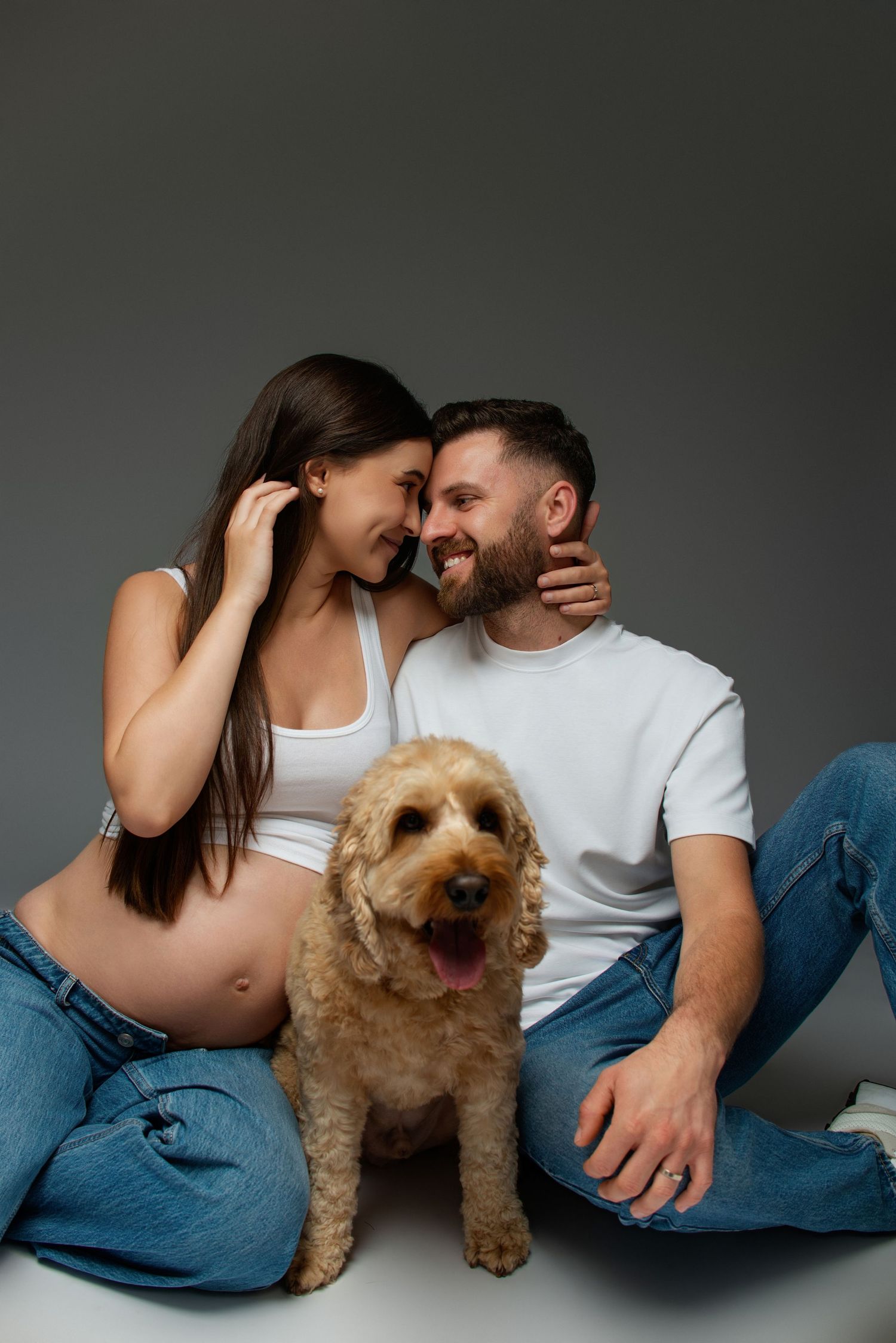 Happy pregnant couple sitting together with a golden doodle dog, smiling intimately in a studio setting.